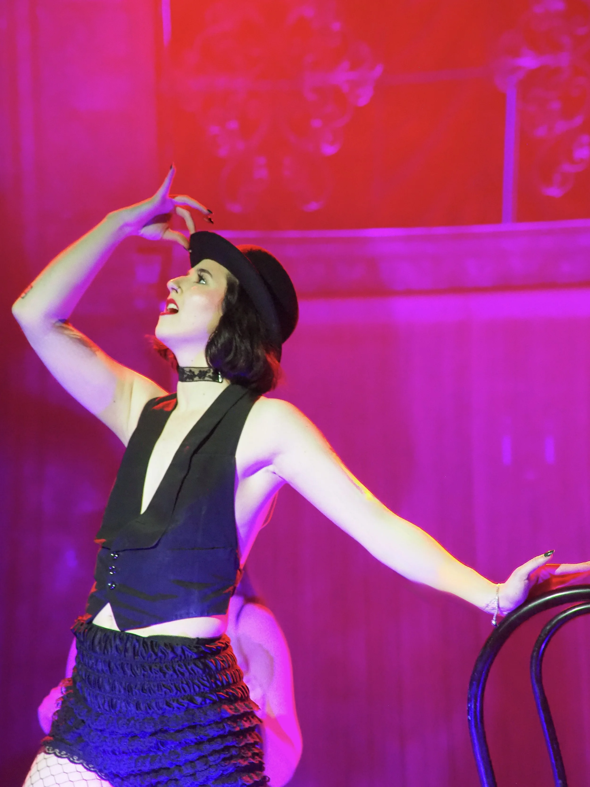 Performer in black outfit, hat, and fishnet stockings on stage with pink lighting.