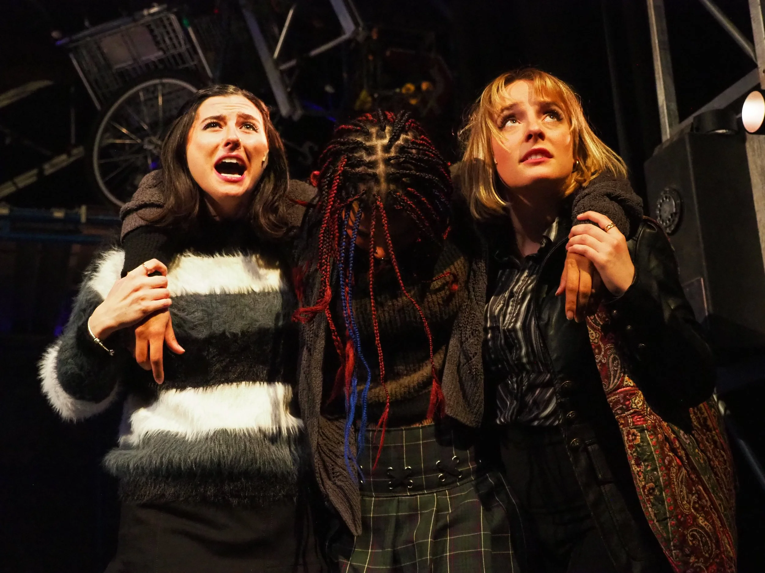 Three women in dressy attire appear distressed, with one woman in the middle hiding her face with her arm, while the other two clutch her and look worried or scared. They are in a dark environment with metal structures and bicycles overhead, resembling a scene from a play or a dramatic moment.