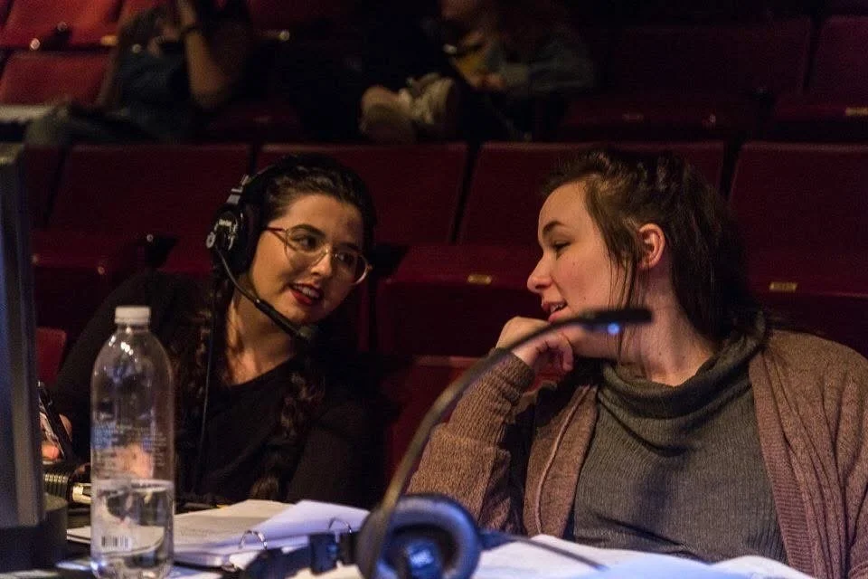 Two women are sitting at a table in a theater or auditorium, engaged in conversation. One woman is wearing a headset and glasses, and the other has her hand resting near her chin. There is a water bottle and papers on the table.