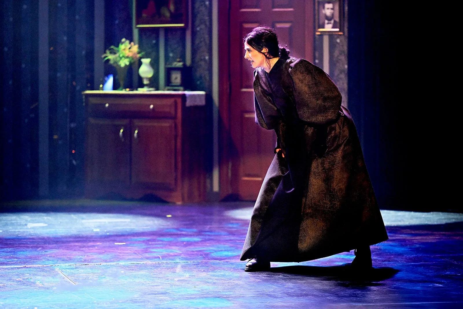 A woman dressed in a long, dark gown is bent over, appearing to be in a dramatic pose, on a stage with wooden flooring and a dark, decorated background featuring a cabinet, a vase with flowers, and framed pictures.