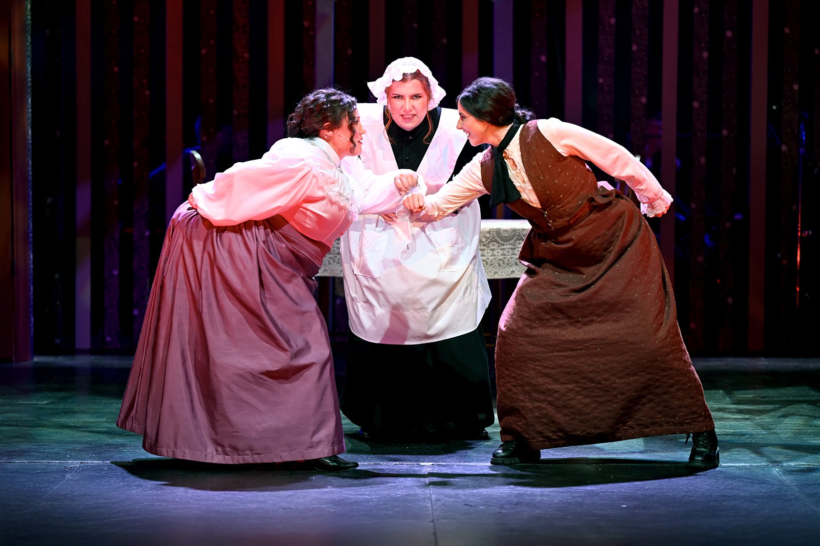 Three women dressed in historical clothing on stage, engaging in a theatrical performance with a nurse in the center. Two women are facing each other, arm wrestling, while the nurse in the middle looks on with a stern expression.