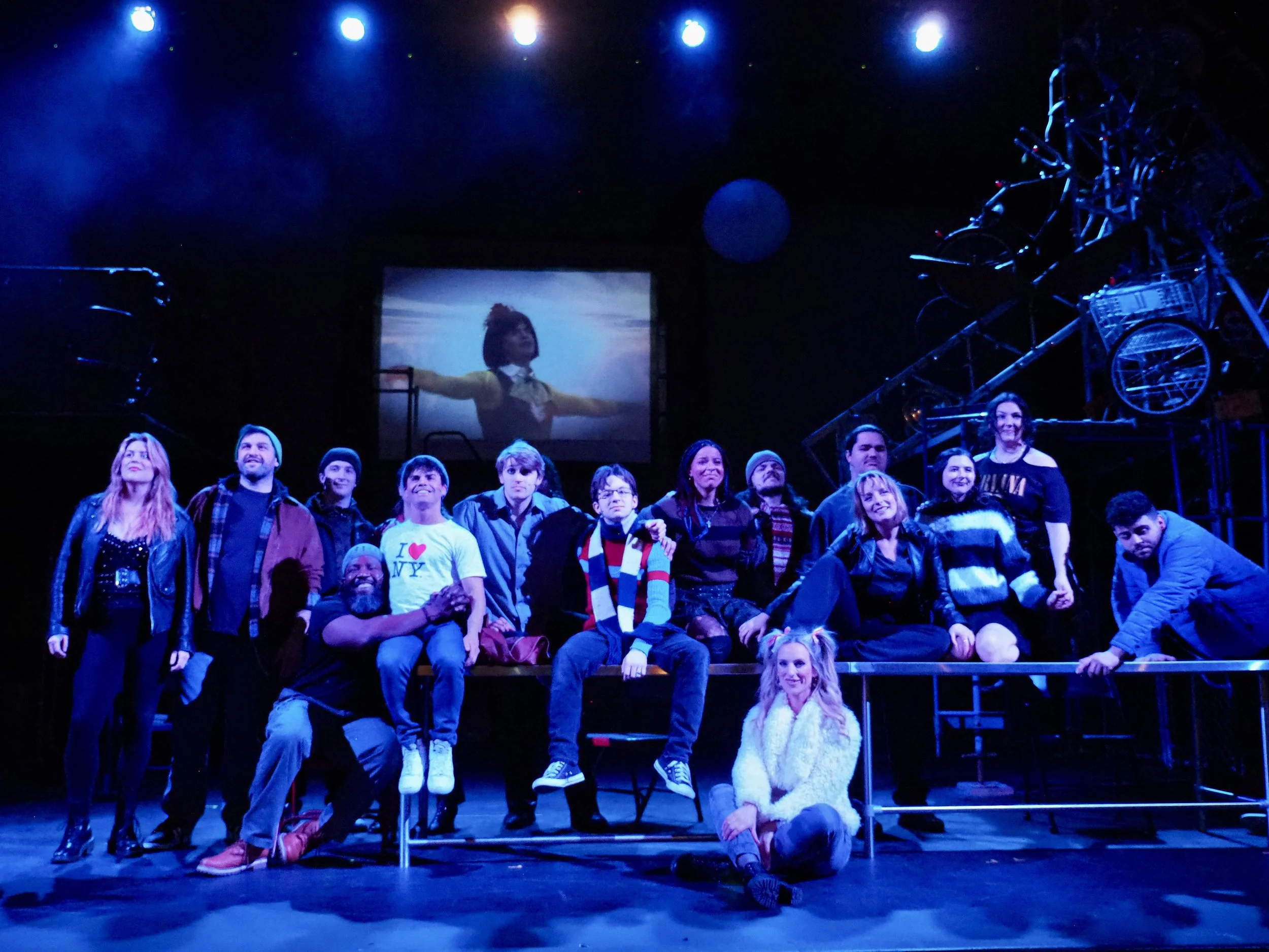 Group of people on stage during a theatrical performance, with a projection of a girl in the background and stage lighting.