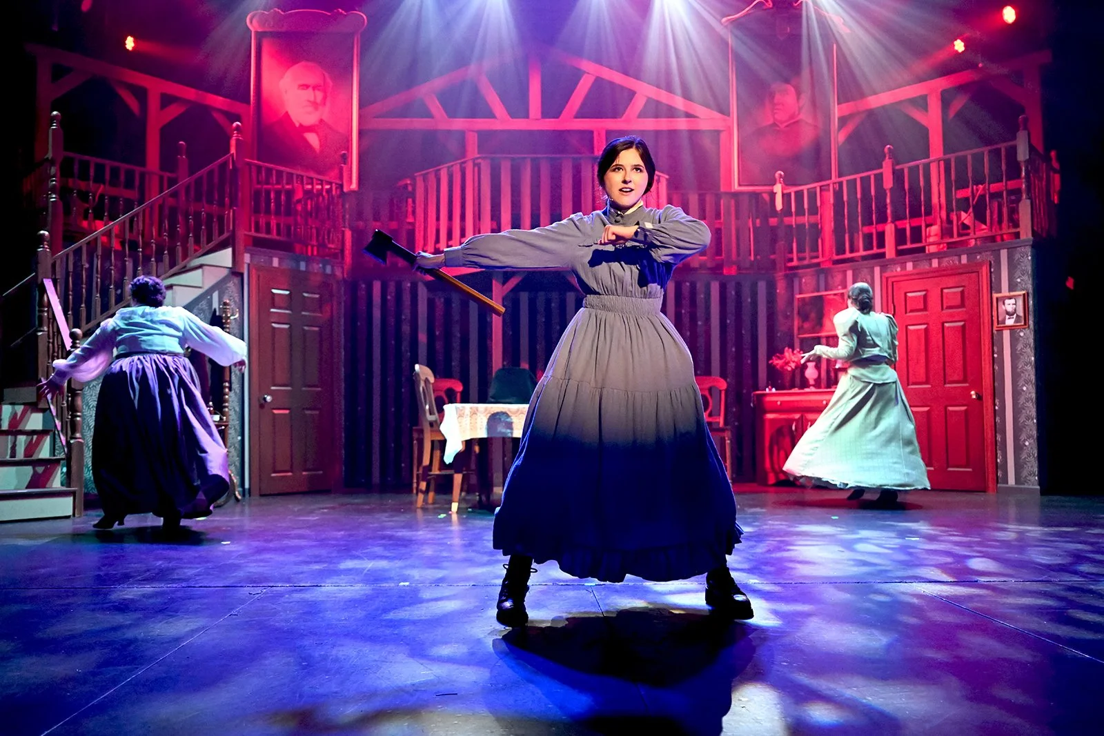 A woman on stage dressed in historical costume, holding an axe, performing in a theatrical play with two other women in period dresses and a detailed set resembling a vintage house interior, with portraits hanging on the wall and theatrical lighting.