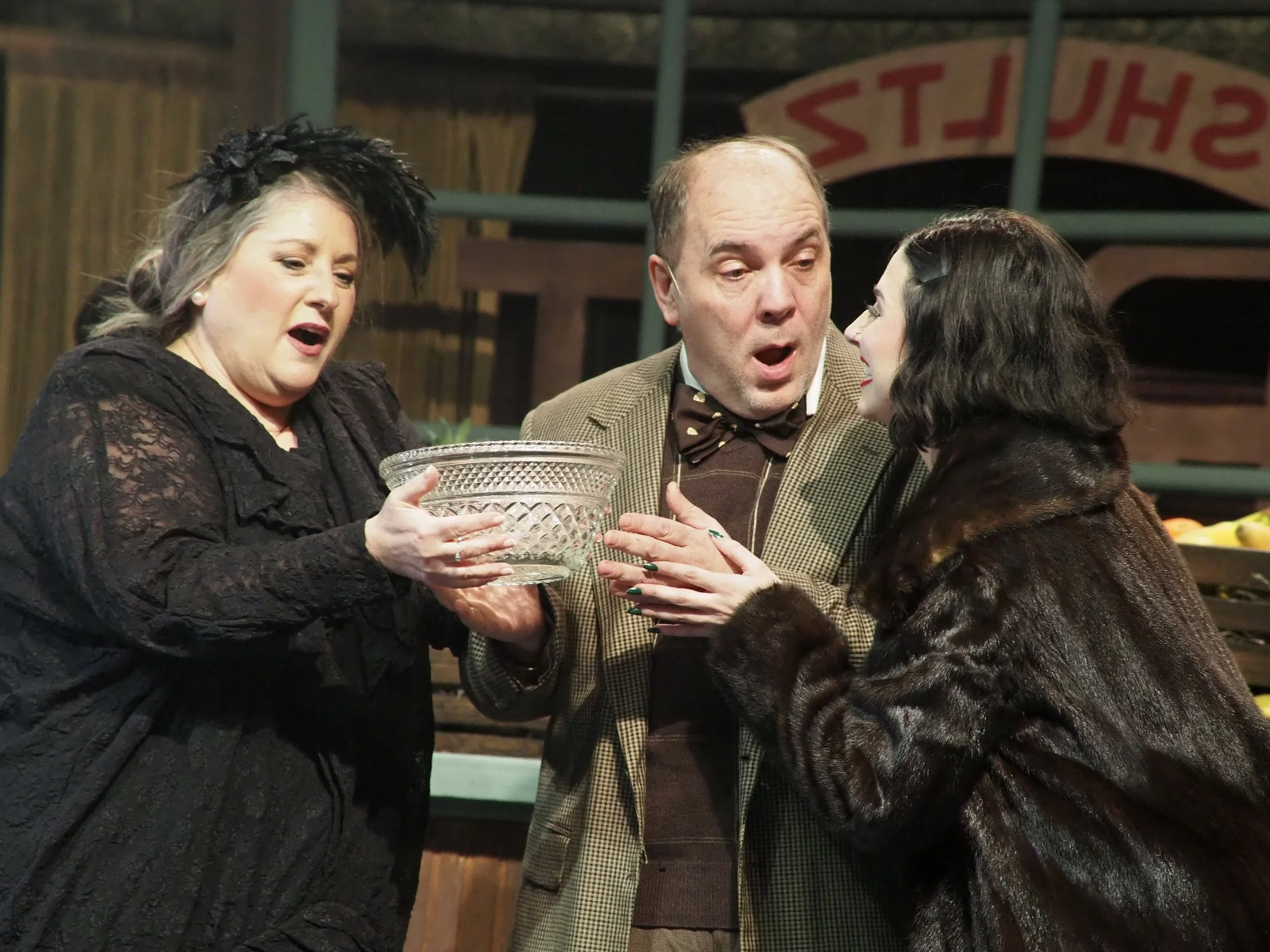 Three actors in a theatrical scene: a woman with gray hair and black clothing holding a glass dish, a man in a brown blazer and bow tie with a surprised expression, and a woman with black hair and a fur coat engaging in a conversation, set against a vintage-style background.