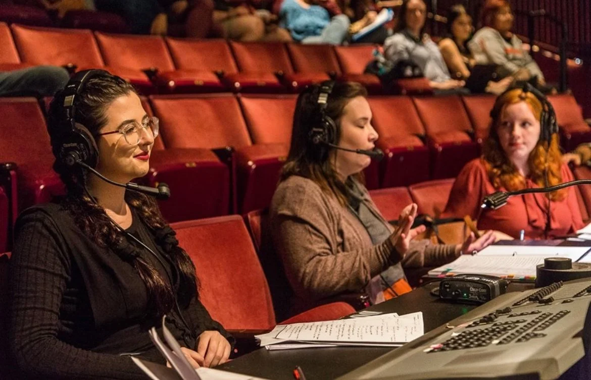 Three women with headsets working as audio or video control operators in a theater or auditorium with red seats and audience in the background.