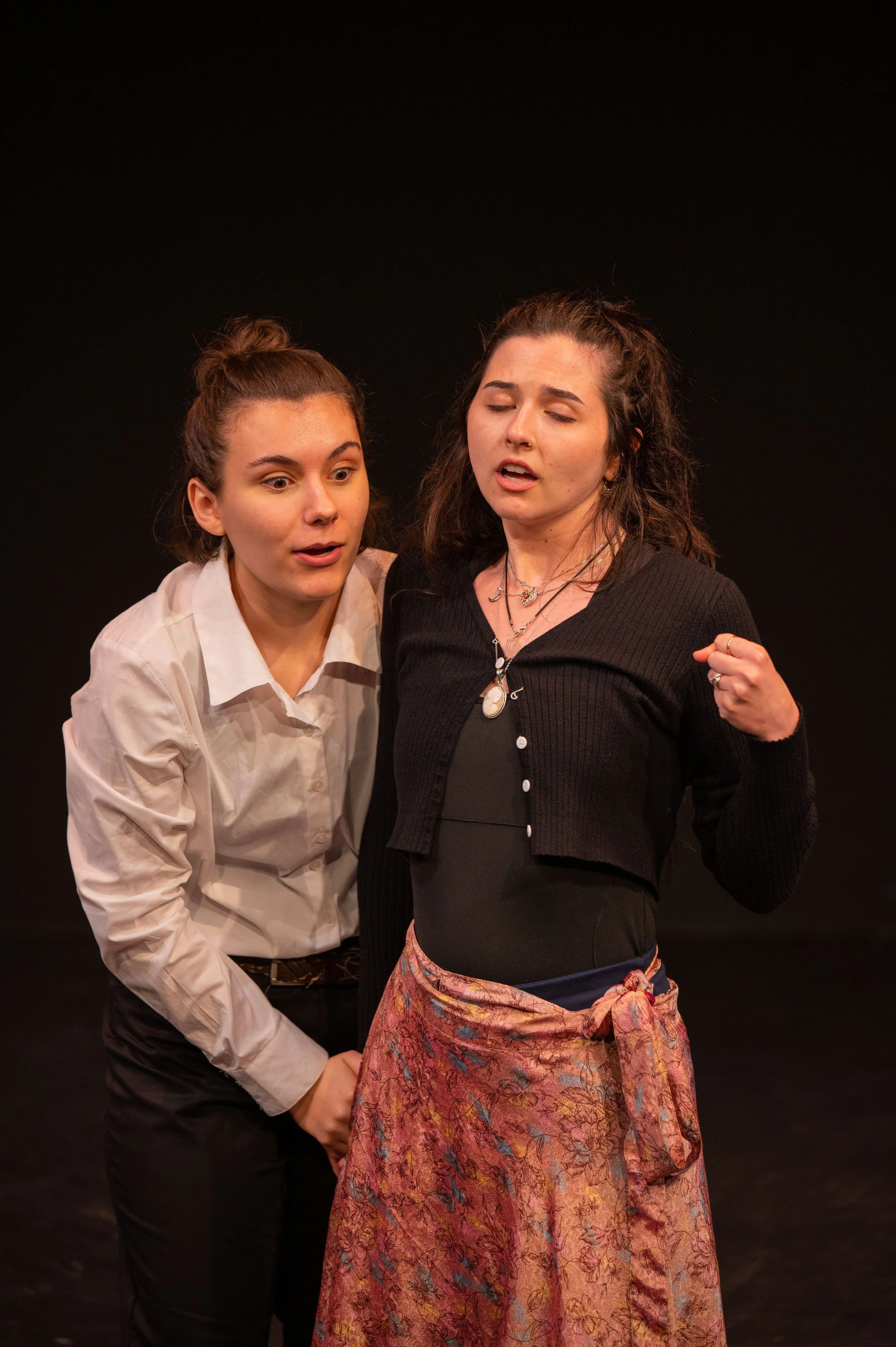 Two women on stage in an intense conversation. The woman on the left, dressed in a white shirt, appears surprised or concerned. The woman on the right, with dark hair and a black cardigan, has a determined expression and clenched fist.