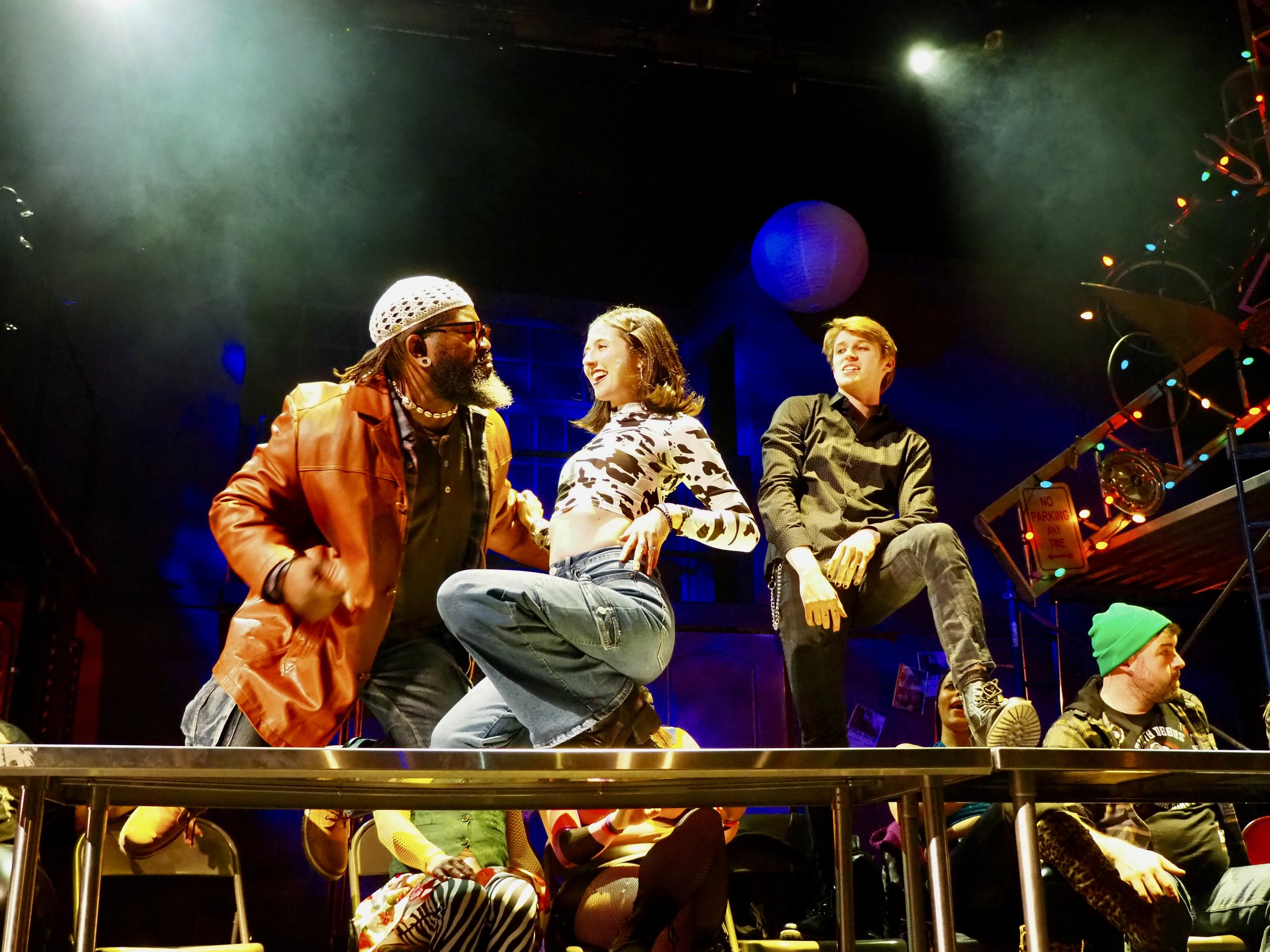 Three people on stage, two men and one woman, appear to be engaged in a lively conversation or performance, with colorful stage lights and decorations around them.