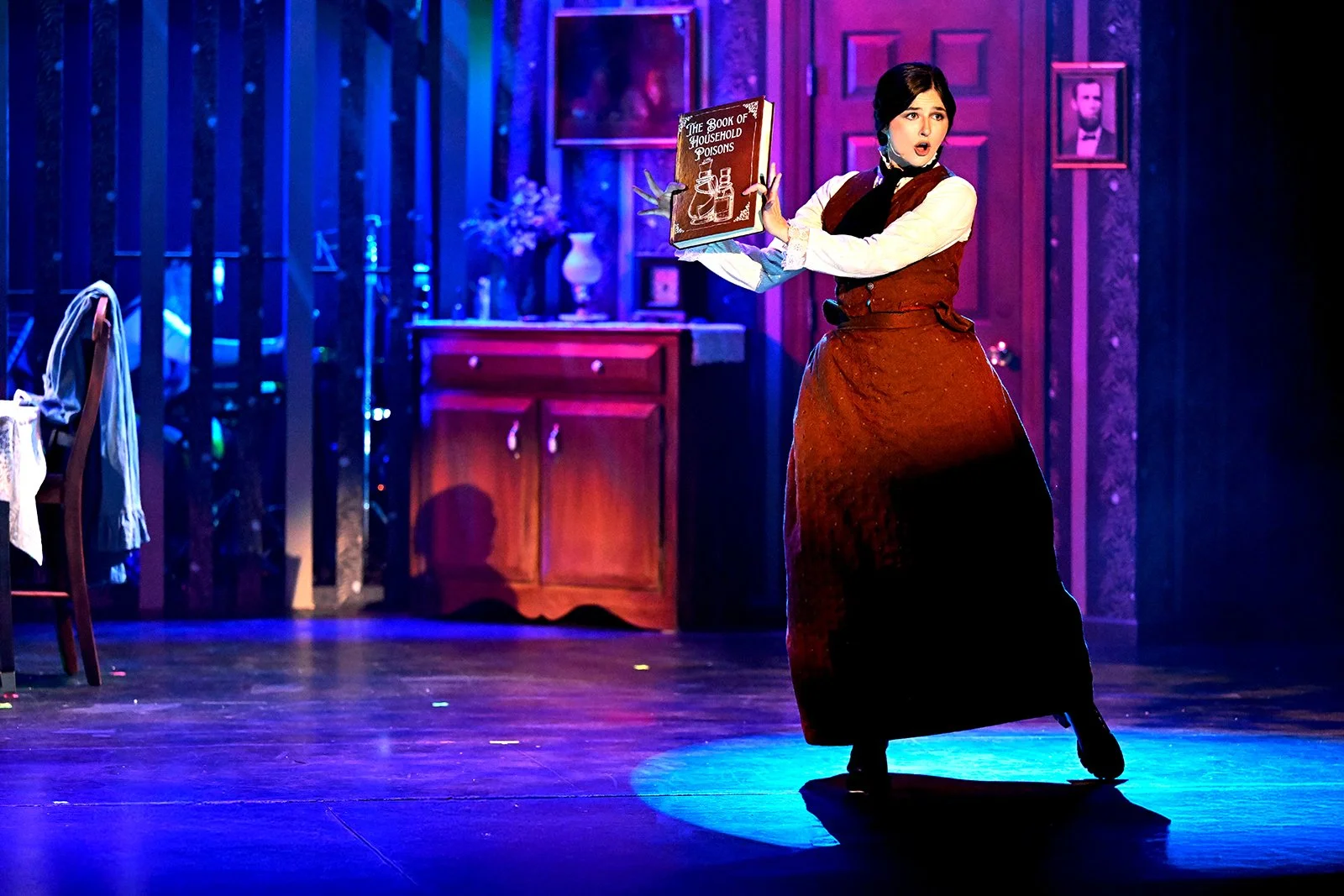 Woman performing on stage, wearing period costume, holding a book titled 'The Book of Household Poisons'. The stage has vintage furniture and framed photos in the background, lit with colorful stage lights.