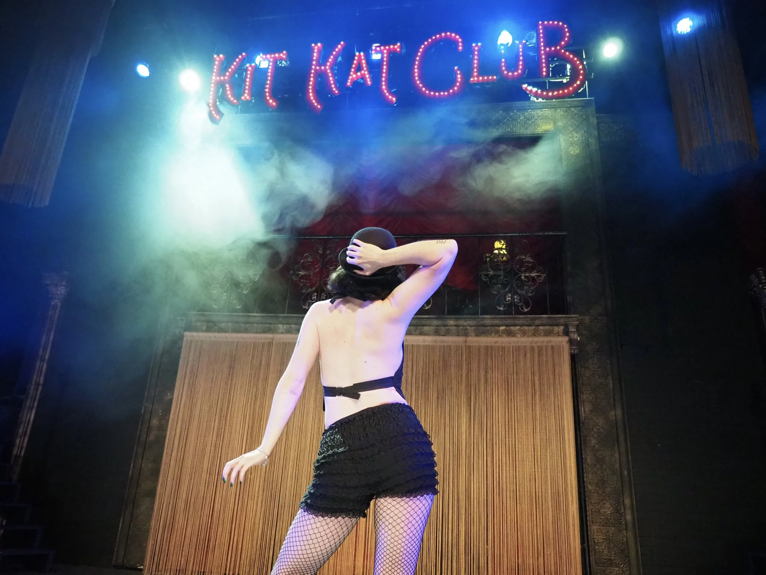 A woman with black hair, wearing fishnet stockings, a black hat, a black top, and a black skirt, stands on a stage in front of a neon sign that reads 'KITT KAT CLUB' amid colorful stage lighting and smoke effects.