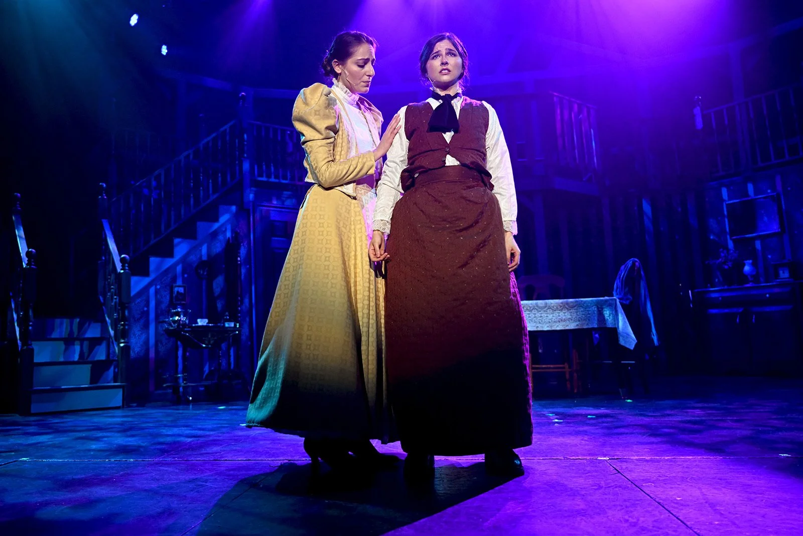 Two women in historical costumes standing on stage with purple lighting, one comforting the other, in a theatrical scene.