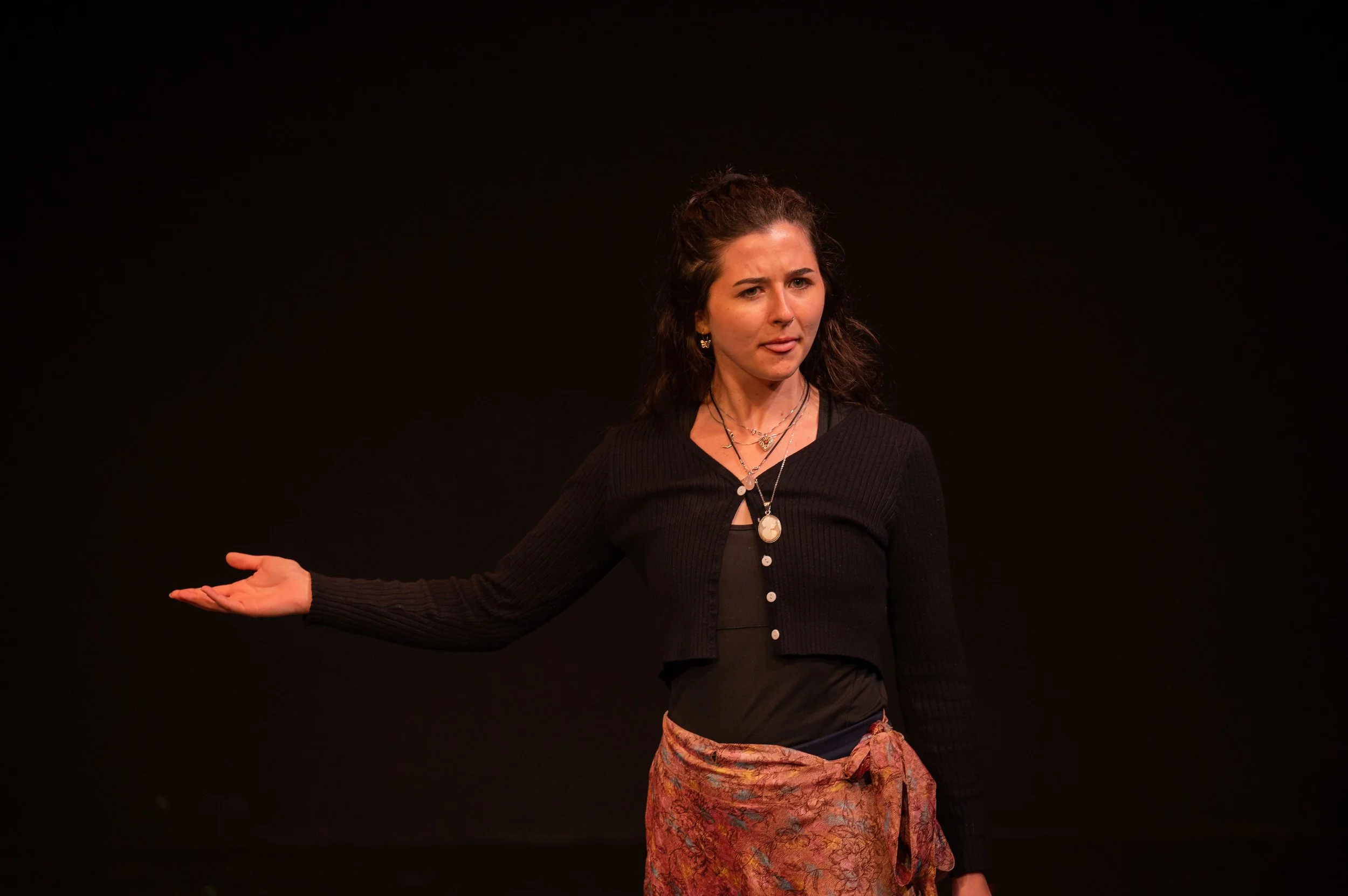A woman with dark curly hair gesturing with her left hand, wearing a black cardigan over a black top, a floral skirt, and layered necklaces, against a black background.