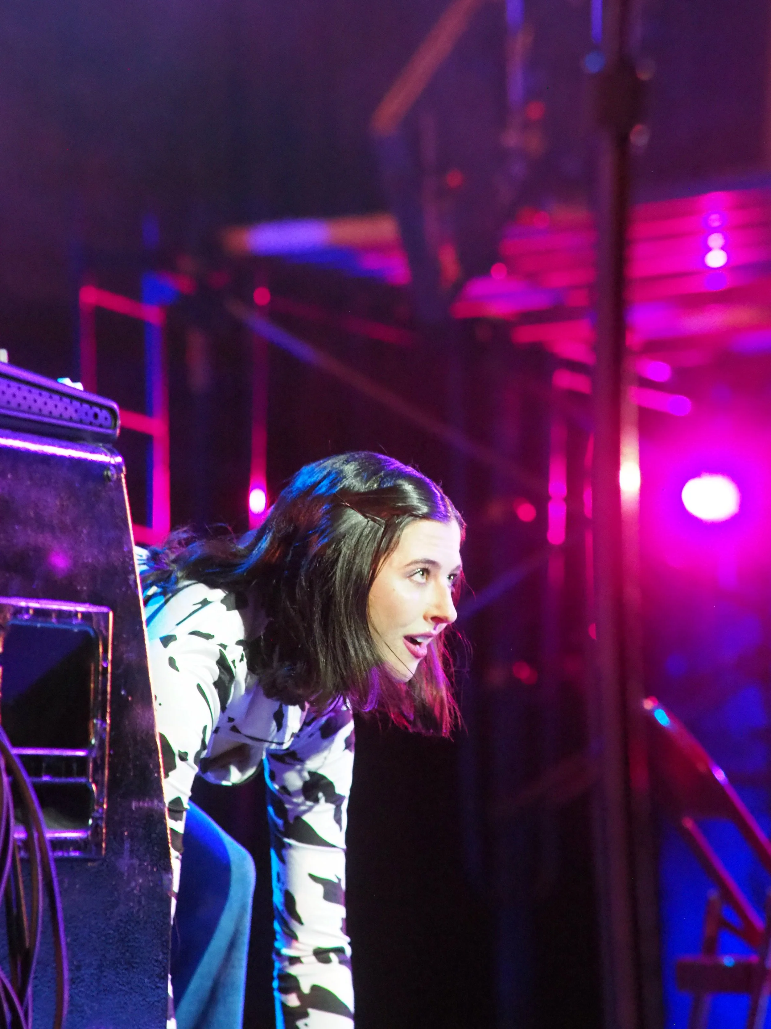 A woman leaning over a turntable or DJ equipment at a concert or nightclub, with colorful stage lighting and purple hues in the background.