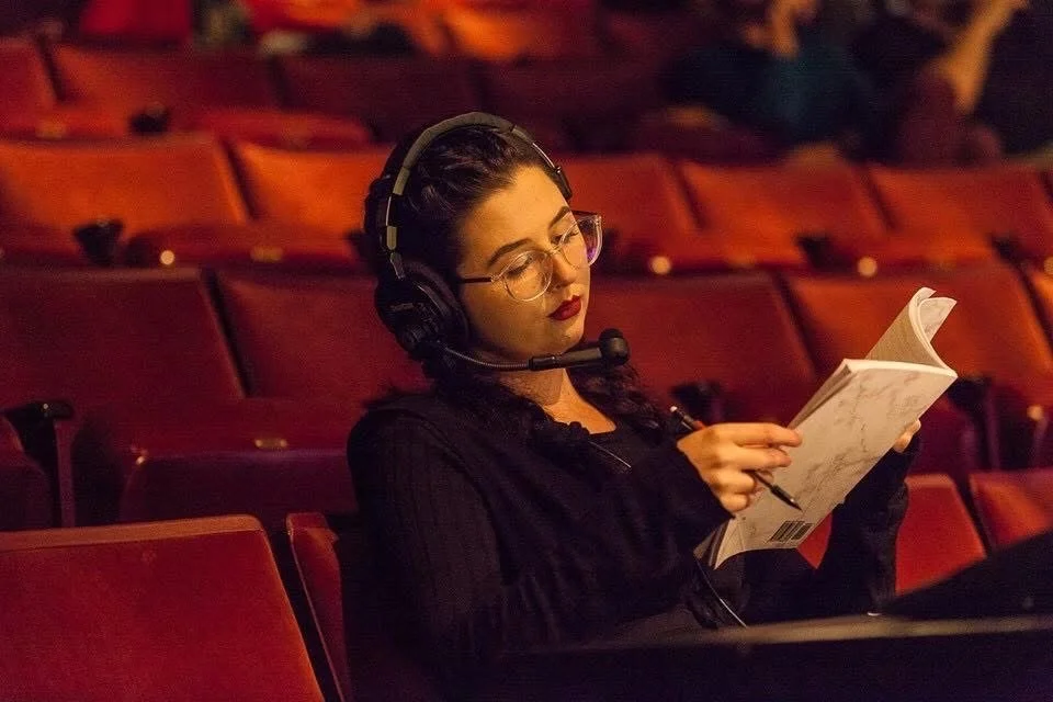 A woman with dark hair wearing glasses and a headset, sitting alone in a theater with red seats, reading a script or booklet.