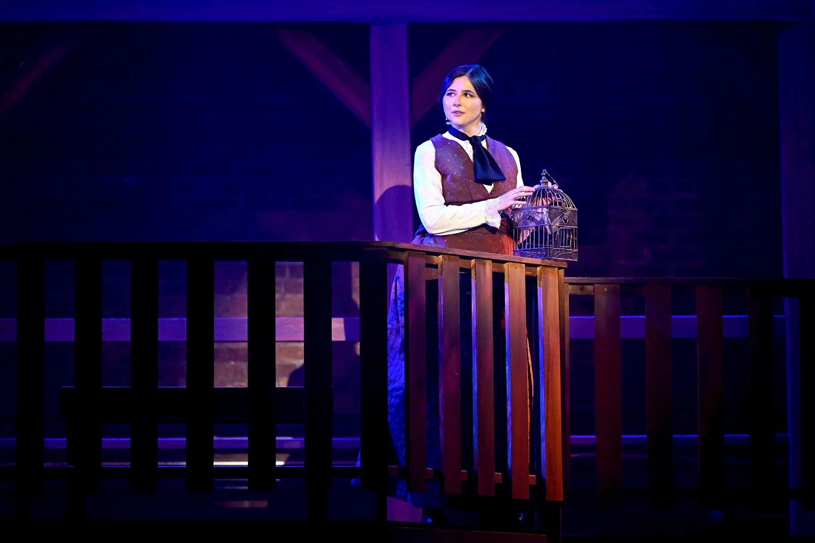 A woman dressed in 19th-century attire holding a birdcage on a wooden balcony inside a theater.