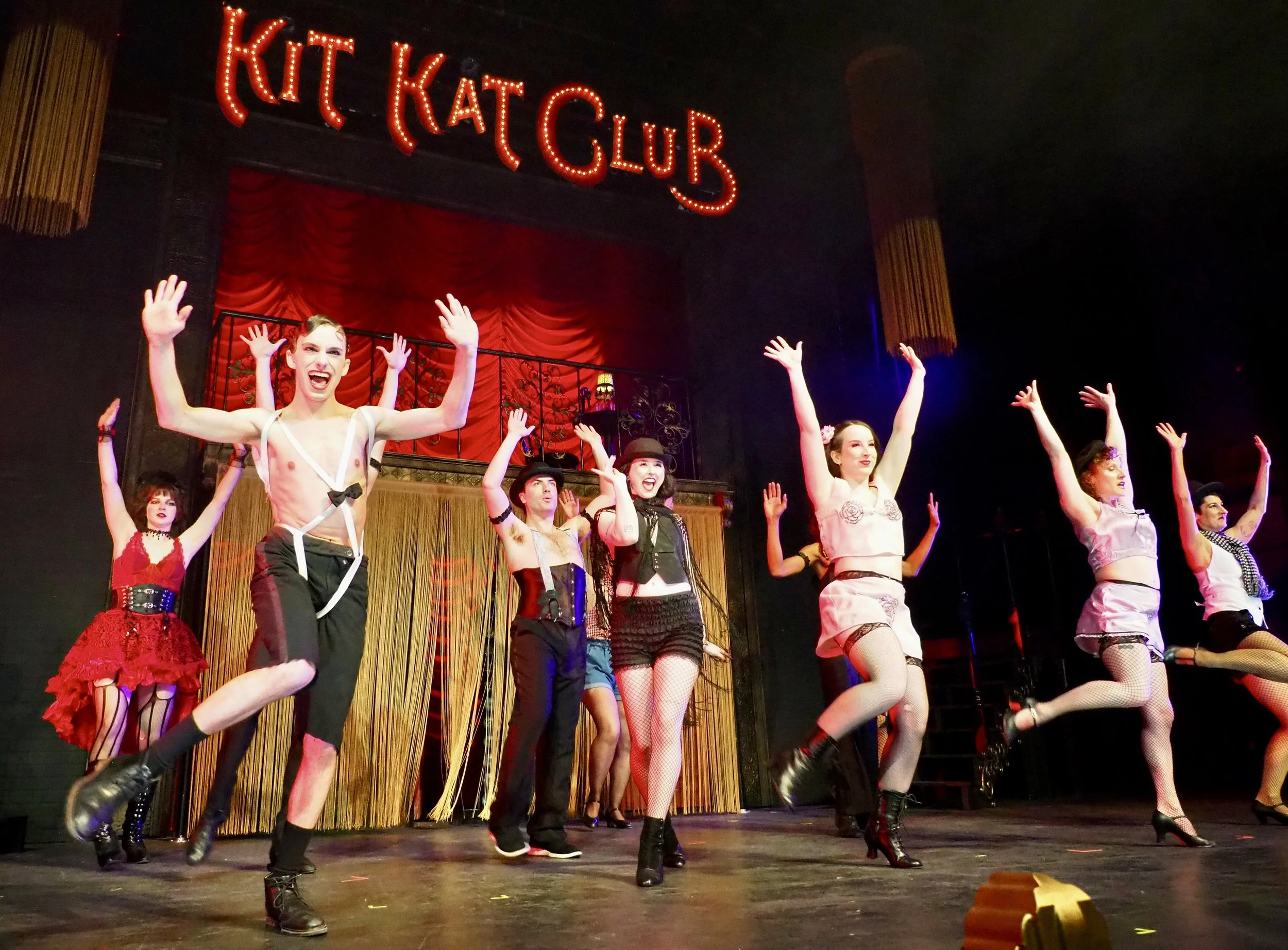 Performers dressed in vintage costumes perform a dance on stage with a red curtain and a sign that reads 'Kit Kat Club' above.