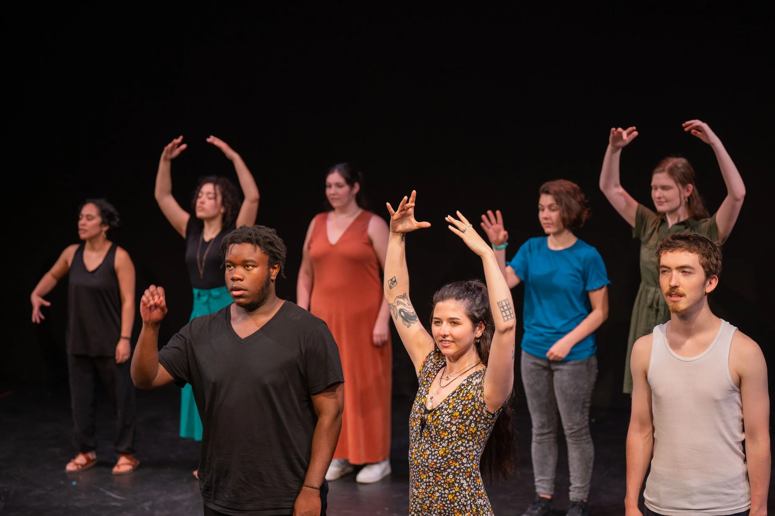 A diverse group of eight people practicing theater or dance on a black stage, with some raising their arms and others standing with relaxed postures.