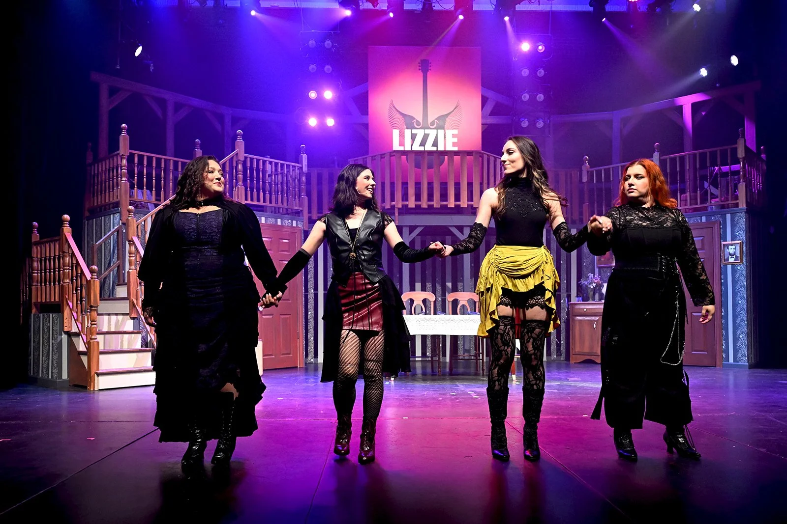 Four women holding hands on stage in a theatrical performance, with a set resembling a house interior in the background and purple stage lighting. A sign with the word 'LIZZIE' and a guitar with wings is displayed above.