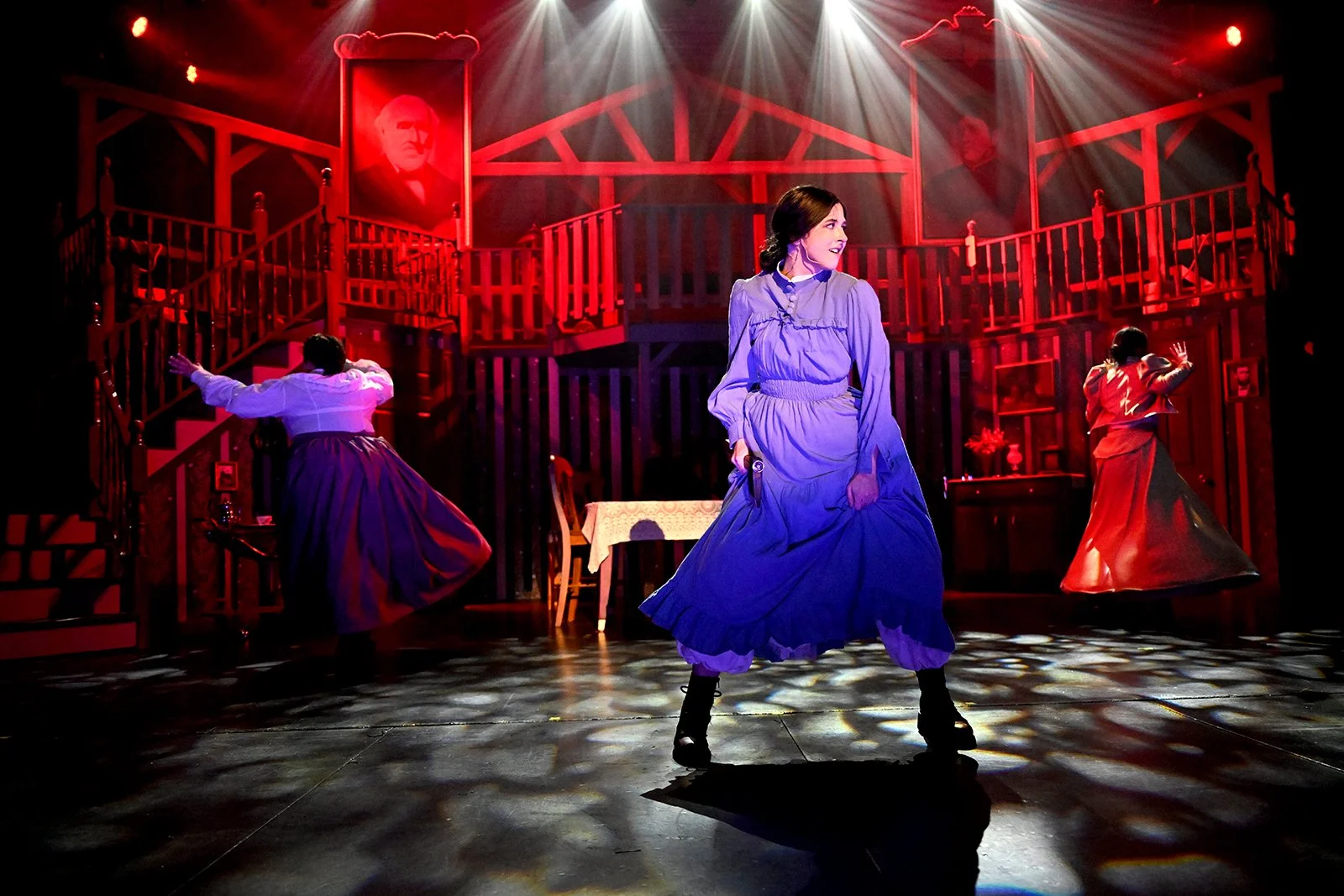 Three women in vintage dresses are performing a dance on stage with a red, wooden two-story interior set, including framed portraits and furniture. Bright stage lights shine overhead.