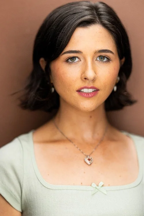 A young woman with short dark hair and blue eyes, wearing a light green top with a small bow detail, a necklace with a heart-shaped pendant, and earrings, standing against a brown background.