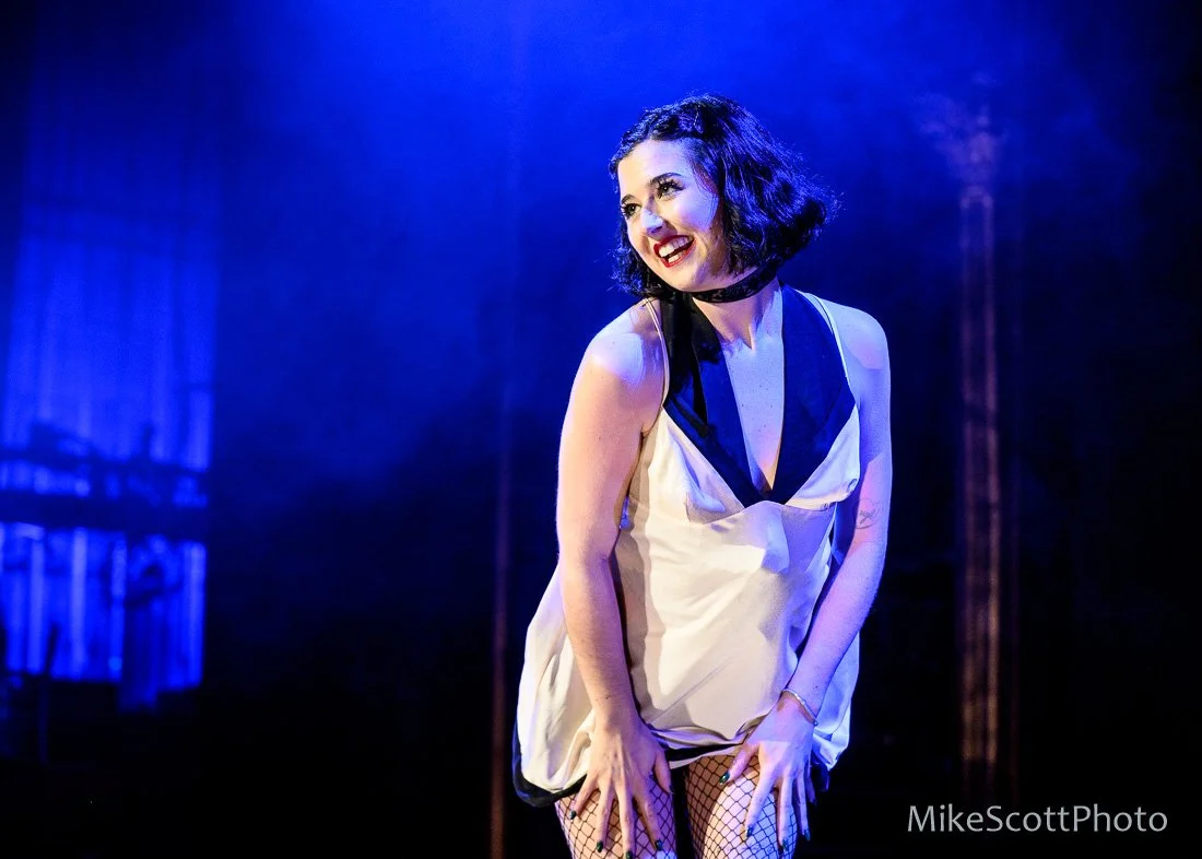 A woman with short black curly hair and red lipstick is performing on stage, wearing a sleeveless beige and black dress with fishnet stockings, under blue stage lighting.