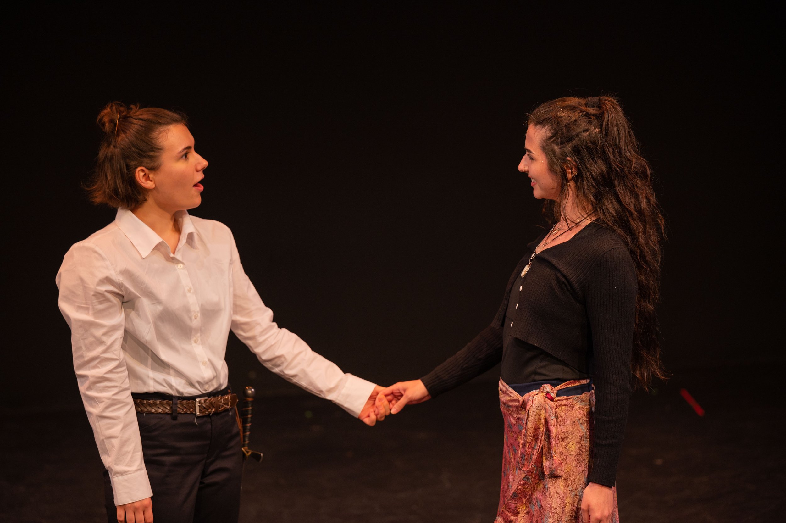 Two women are holding hands and facing each other against a black background. The woman on the left has short brown hair in a bun, is wearing a white shirt and dark pants, and appears surprised. The woman on the right has long wavy brown hair, is wearing a black top and patterned pants, and is smiling.