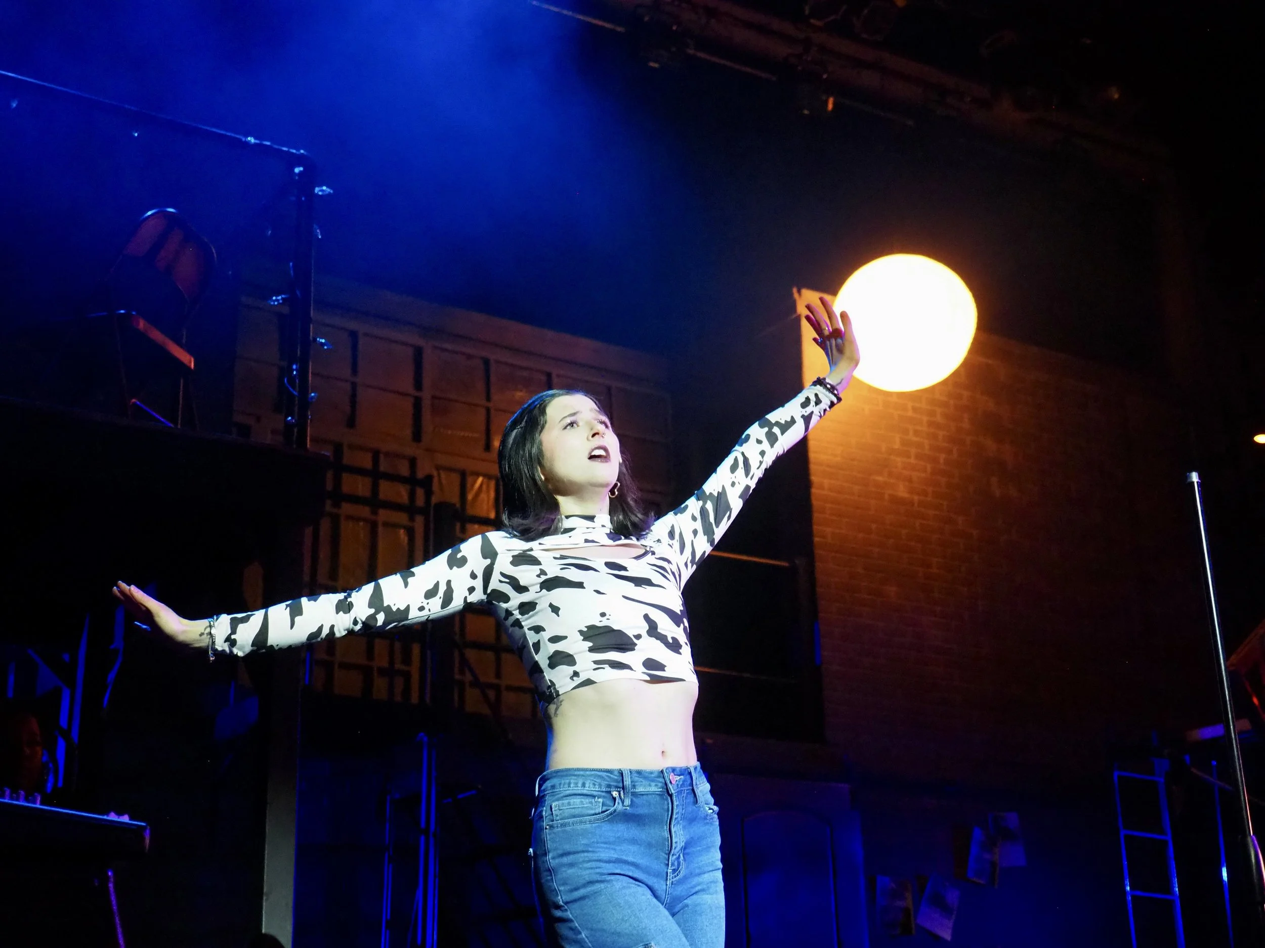 A woman with black hair in a cropped, long-sleeve shirt with a black and white cow print pattern and blue jeans stands on stage with arms outstretched. She appears to be singing or speaking, with a bright light (moon-like orb) in the background and stage equipment visible around her.
