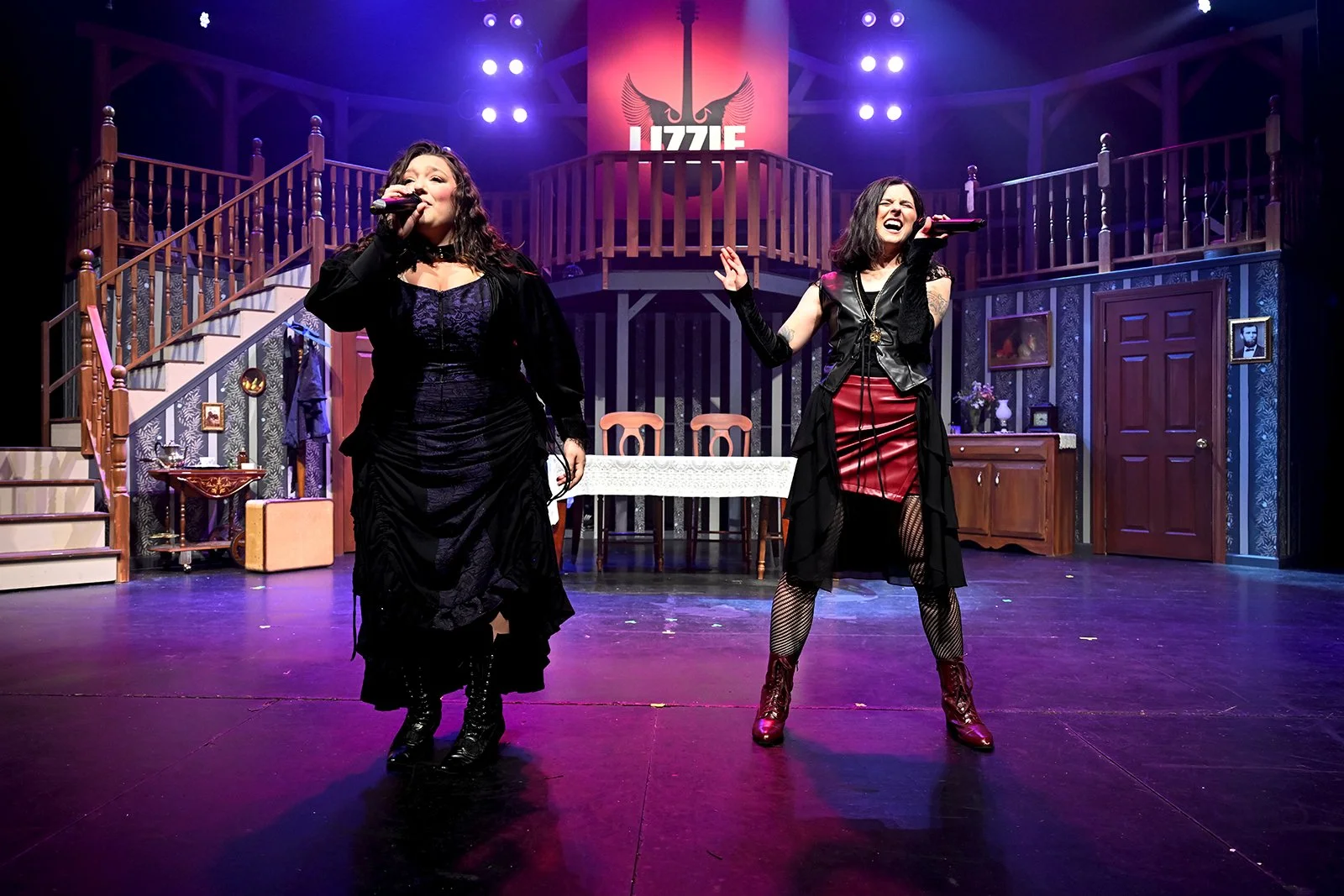 Two women performing on stage in a theater set with a staircase, furniture, and a large sign that reads 'Lizzie' with a black swan logo. One woman, on the left, is wearing a long black dress, and the other, on the right, is in a black top, red skirt, and fishnet stockings, both singing into microphones.