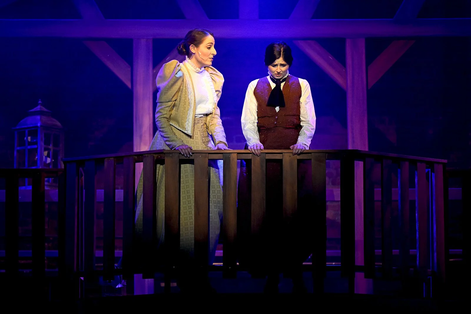 Two women on stage during a theatrical performance, standing behind a wooden railing with a dark background. One woman is dressed in vintage attire with a yellow jacket, while the other wears a vest and a tie.