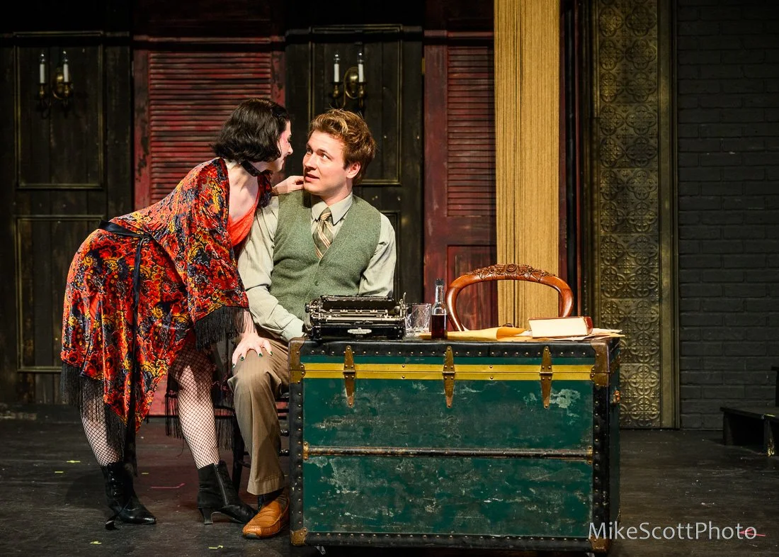 A woman in a red dress and a patterned shawl leans over a seated man at a vintage desk on stage. The man wears a green vest and tie, with a typewriter and drinks on the desk. The scene appears theatrical with dark background and stage lights.