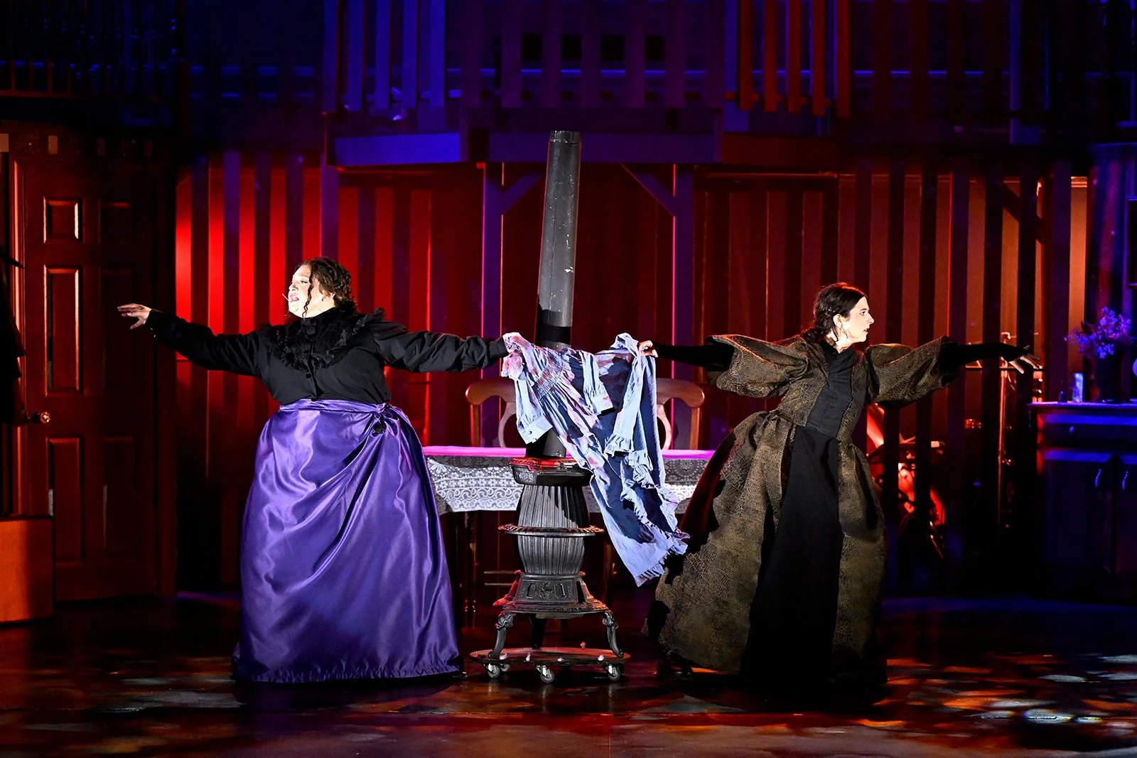 Two women on stage, performing in historical costumes, holding a clothesline with laundry between them, in a theater with red and purple lighting.
