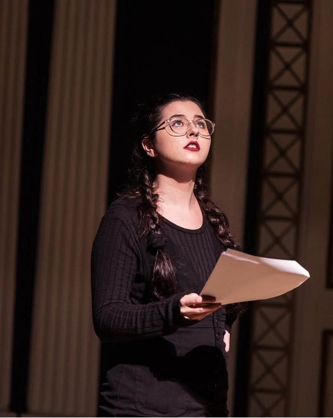 A young woman with glasses, dark hair in braids, wearing a black long-sleeve shirt, holding papers, standing on a stage with curtains and decorative panels in the background.