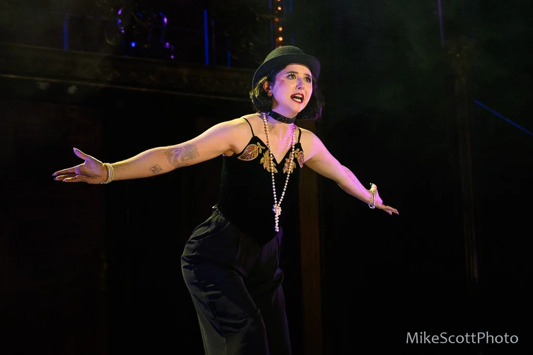 A woman performing on stage, wearing a black top hat, a black sleeveless outfit with gold embellishments, a pearl necklace, and black pants, with an expressive facial expression and arms outstretched.