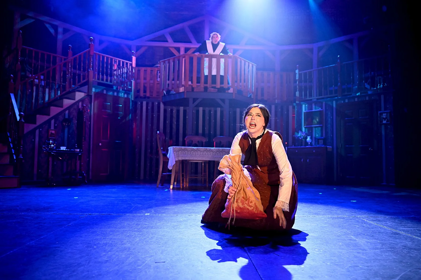 The image shows a theatrical performance with a woman kneeling on the stage, holding a bag, and expressing a passionate emotion. In the background, there is a multi-level wooden set resembling a house interior, with a person standing on the upper balcony observing the scene. The stage is lit with blue and purple lighting, creating a dramatic atmosphere.