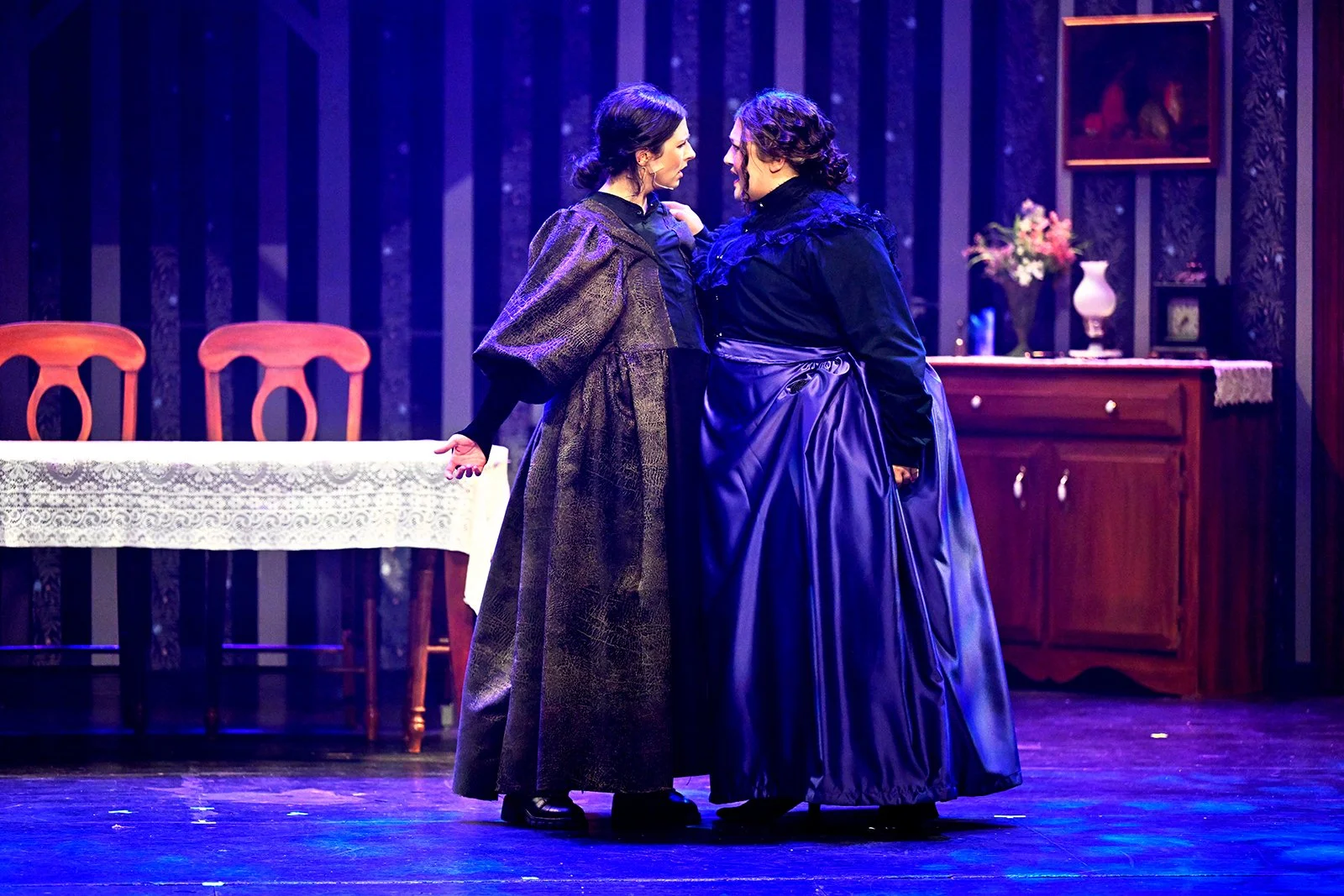 Two women in period costumes standing closely on stage, facing each other, in what appears to be an intense or emotional moment in a theatrical performance. The stage background has a table with a lace tablecloth, chairs, and a wooden sideboard with decorative items and flower arrangements.
