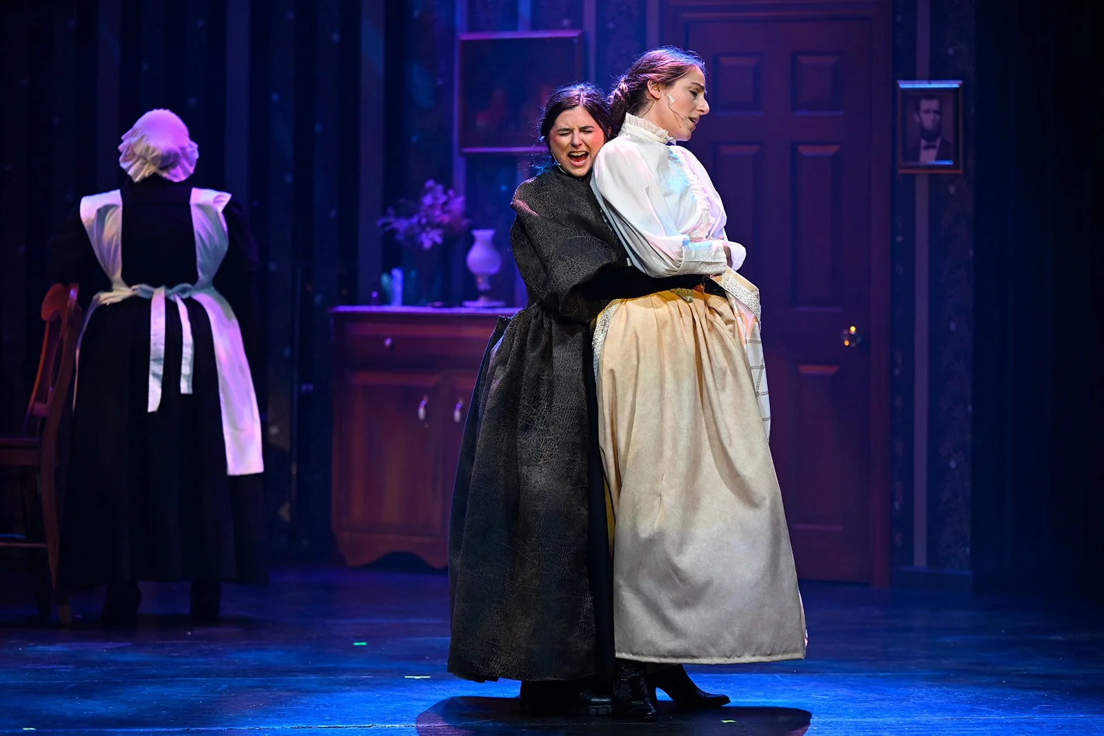Two women are performing on stage, one is crying and the other is comforting her, in period costumes. A maid is seen in the background.