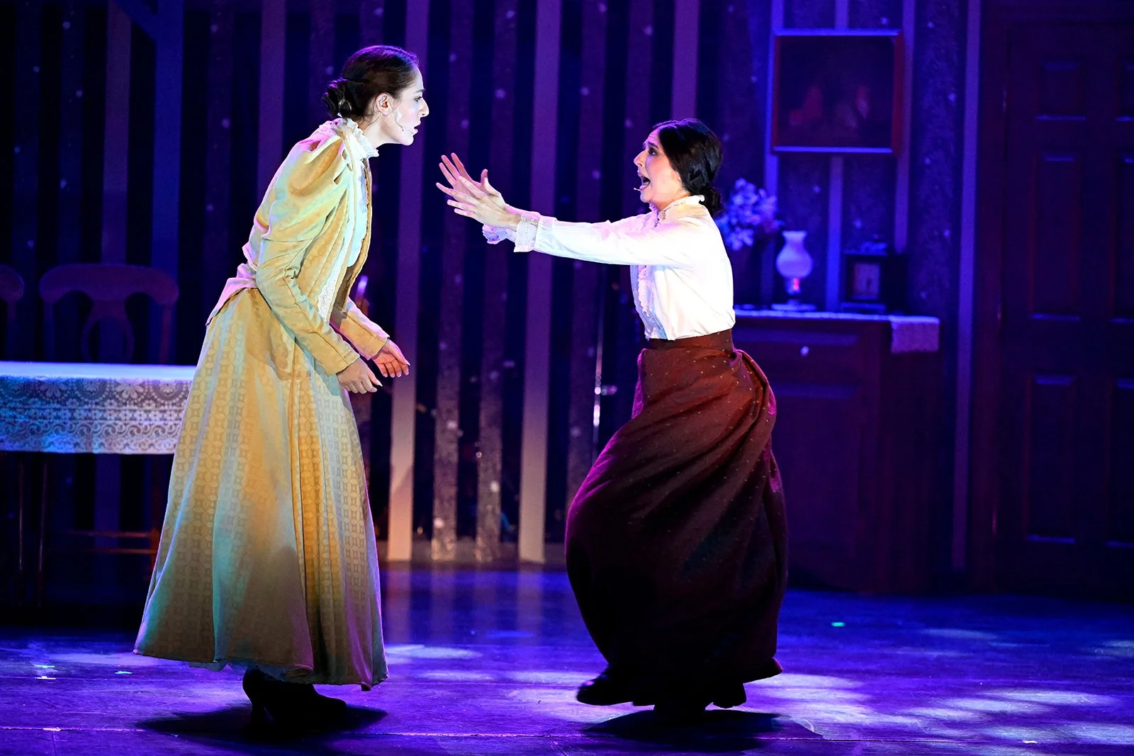 Two women on stage, one in a yellow dress and the other in a white blouse and brown skirt, appear to be engaged in an intense confrontation or argument in a theatrical setting.