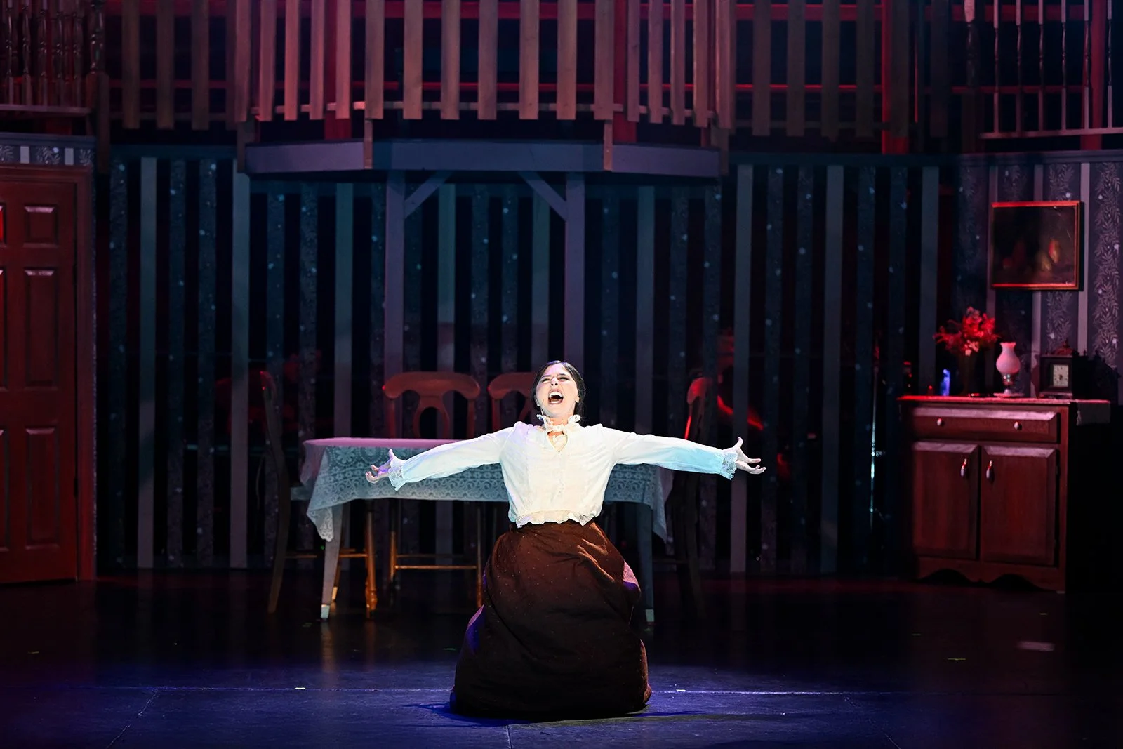 A woman on stage performing with arms outstretched, wearing a white blouse and dark skirt, with a set designed like a cozy, vintage room in the background.