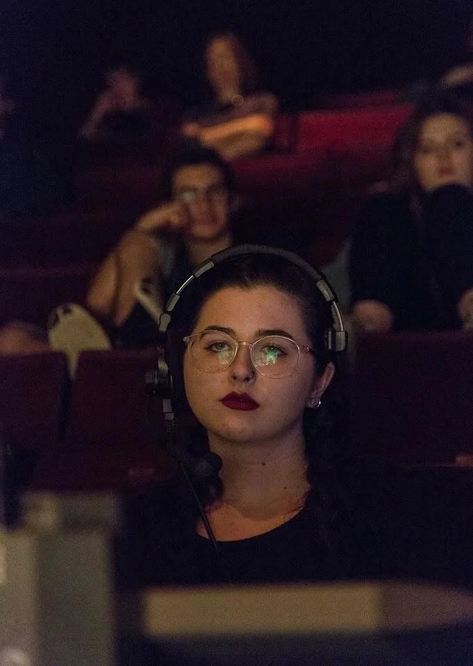 Young woman with dark hair, wearing glasses and red lipstick, listening to headphones during a performance or event in a dark venue.