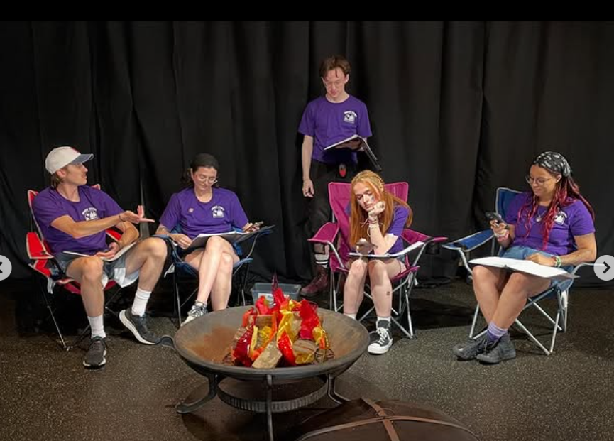 Six young people sitting and standing around a fire pit with decorative flames, all wearing matching purple shirts, in front of a black curtain.