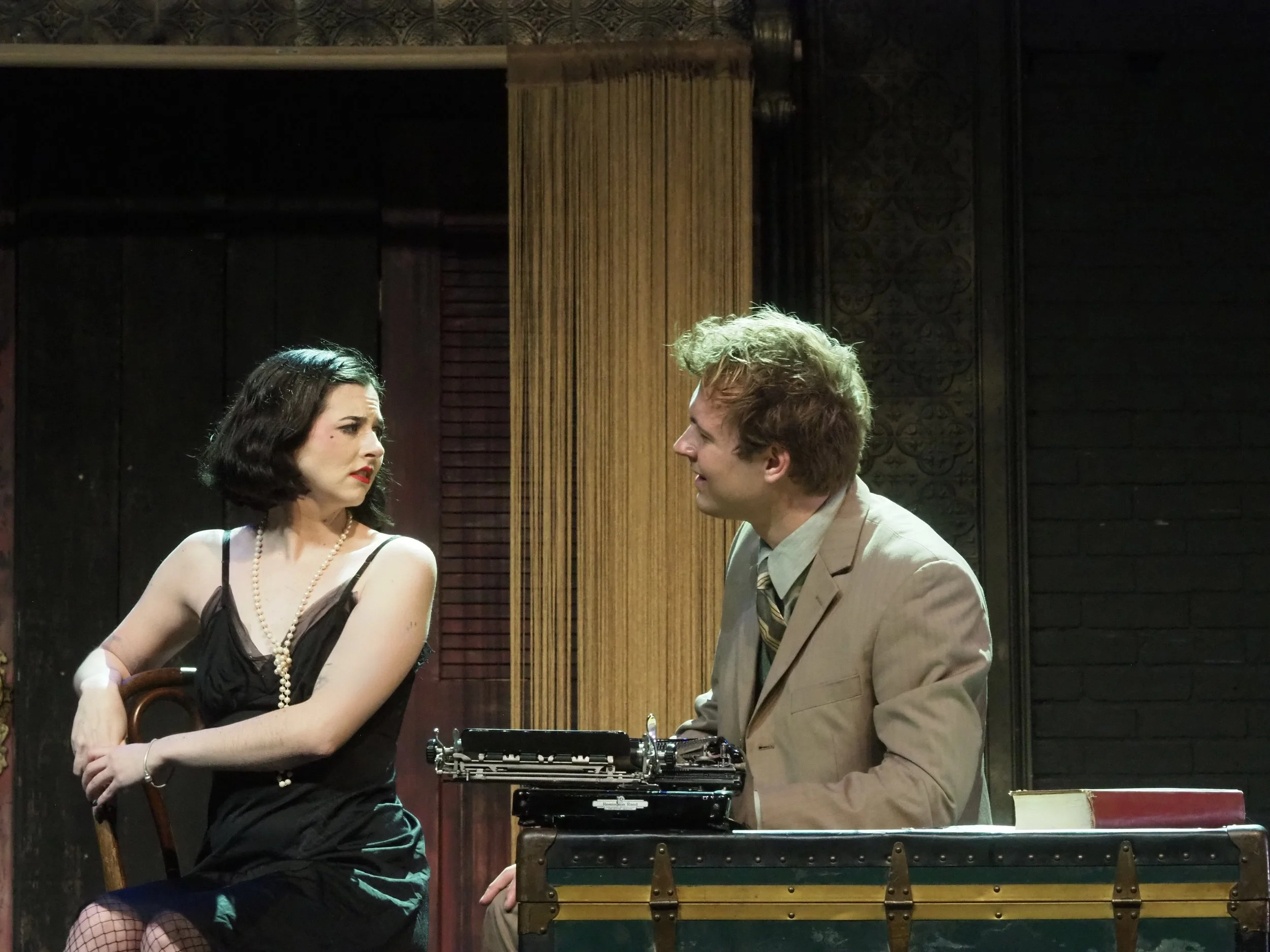 A woman dressed in a black dress and pearls sitting on a chair with arms crossed, looking concerned, while a man in a beige suit and tie sits at a desk with a typewriter, smiling at her, in a dimly lit room with dark wood paneling.