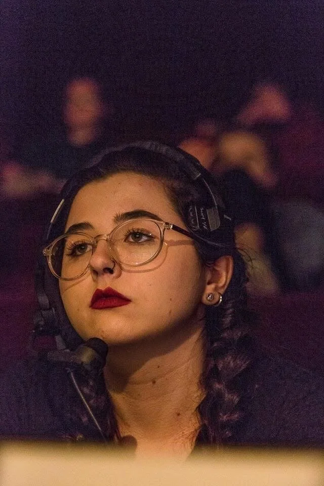 A young woman with glasses and braided dark hair, wearing a headset, in a dark environment, possibly a recording studio or control room.
