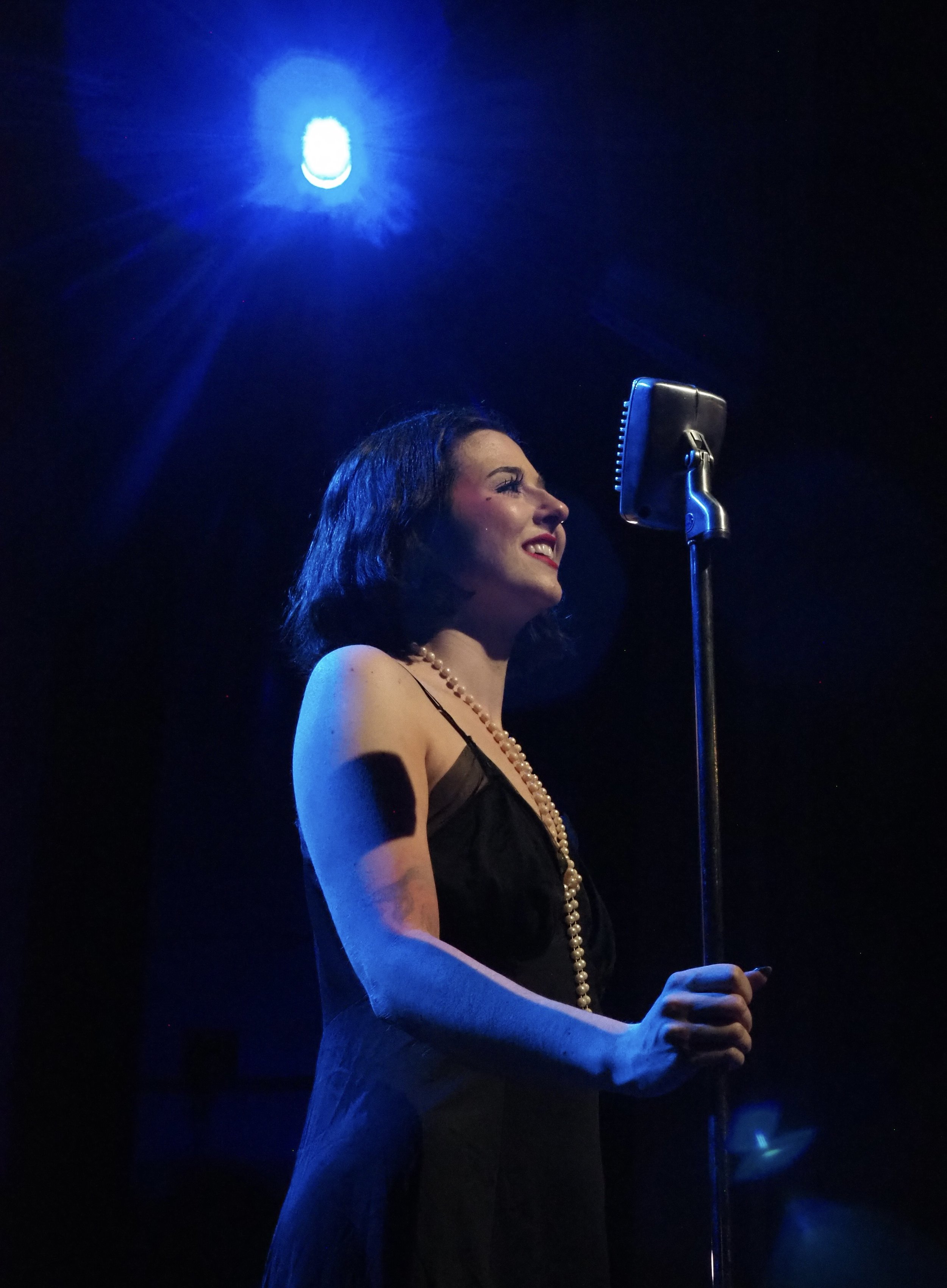 A woman with dark hair, wearing a black dress and pearl necklace, singing into a vintage microphone on stage under blue lighting.