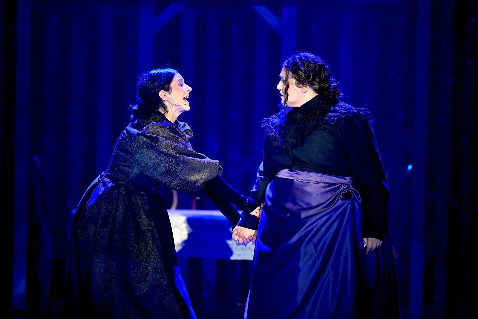 Two women in dark dresses holding hands and engaging in an emotional confrontation on stage with a dark blue background.