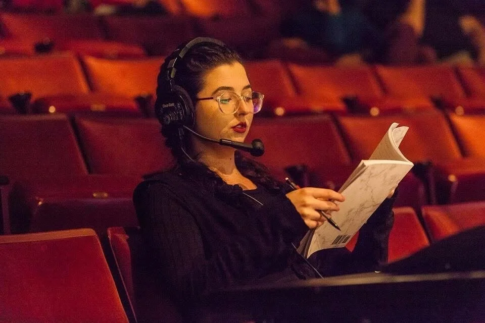 A woman sitting alone in a theater or auditorium, reading a script or booklet, wearing headphones with a microphone.