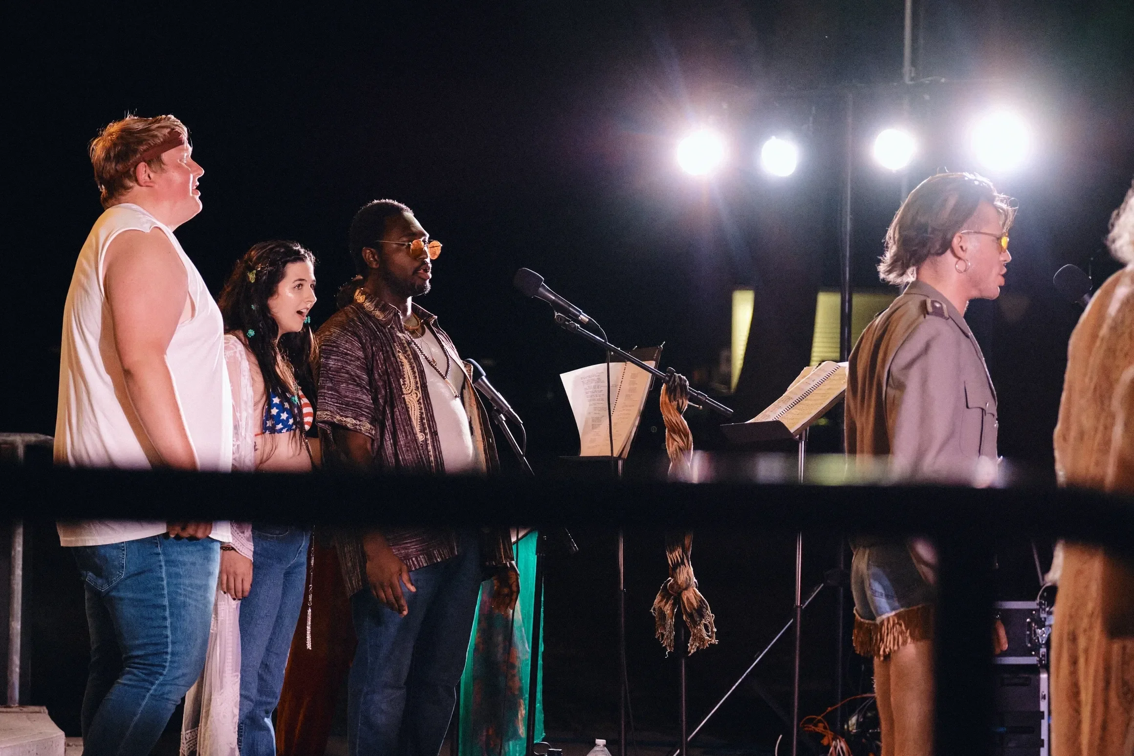 Group of people singing on stage under bright stage lights, with music stands and microphones.
