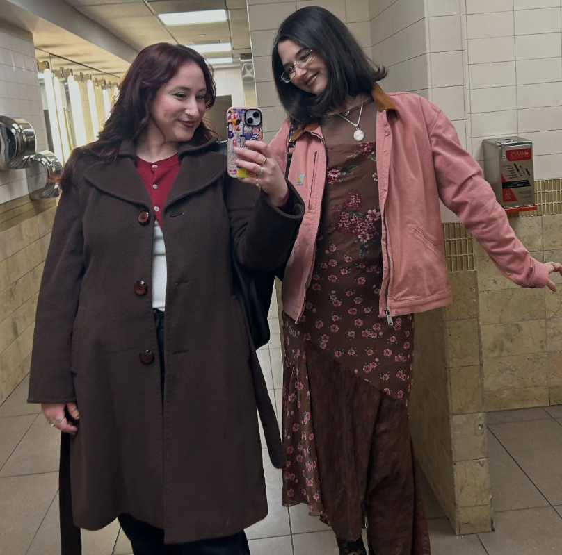 Two women taking a selfie in a public restroom mirror, both smiling. One woman has dark brown hair and is wearing a long brown coat over a white shirt. The other woman has black hair, glasses, and is wearing a pink jacket over a floral dress.