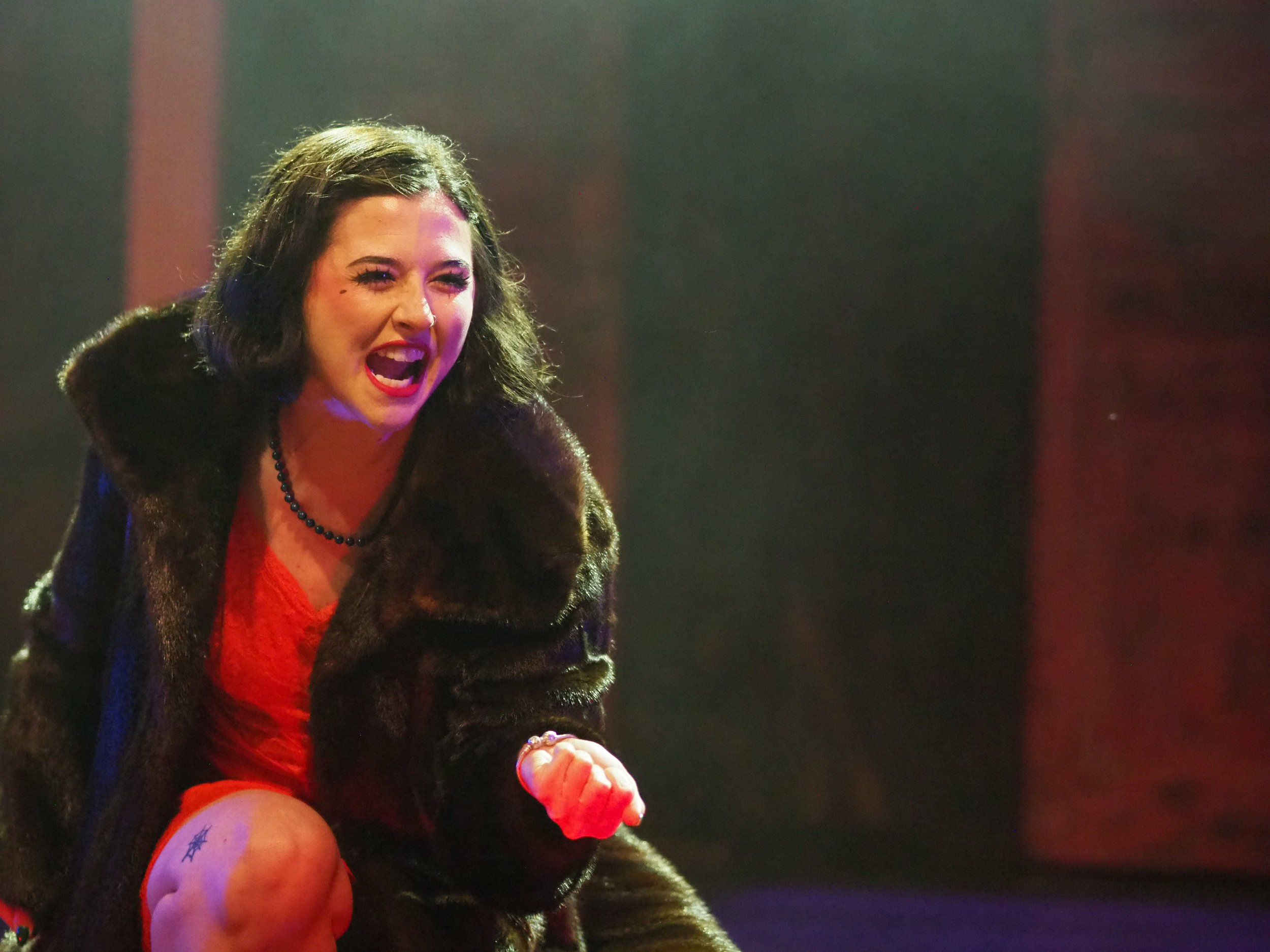 A woman with dark, curly hair expressing intense emotion, wearing a red dress, black necklace, and a black coat with a fur collar, on stage with dramatic lighting.
