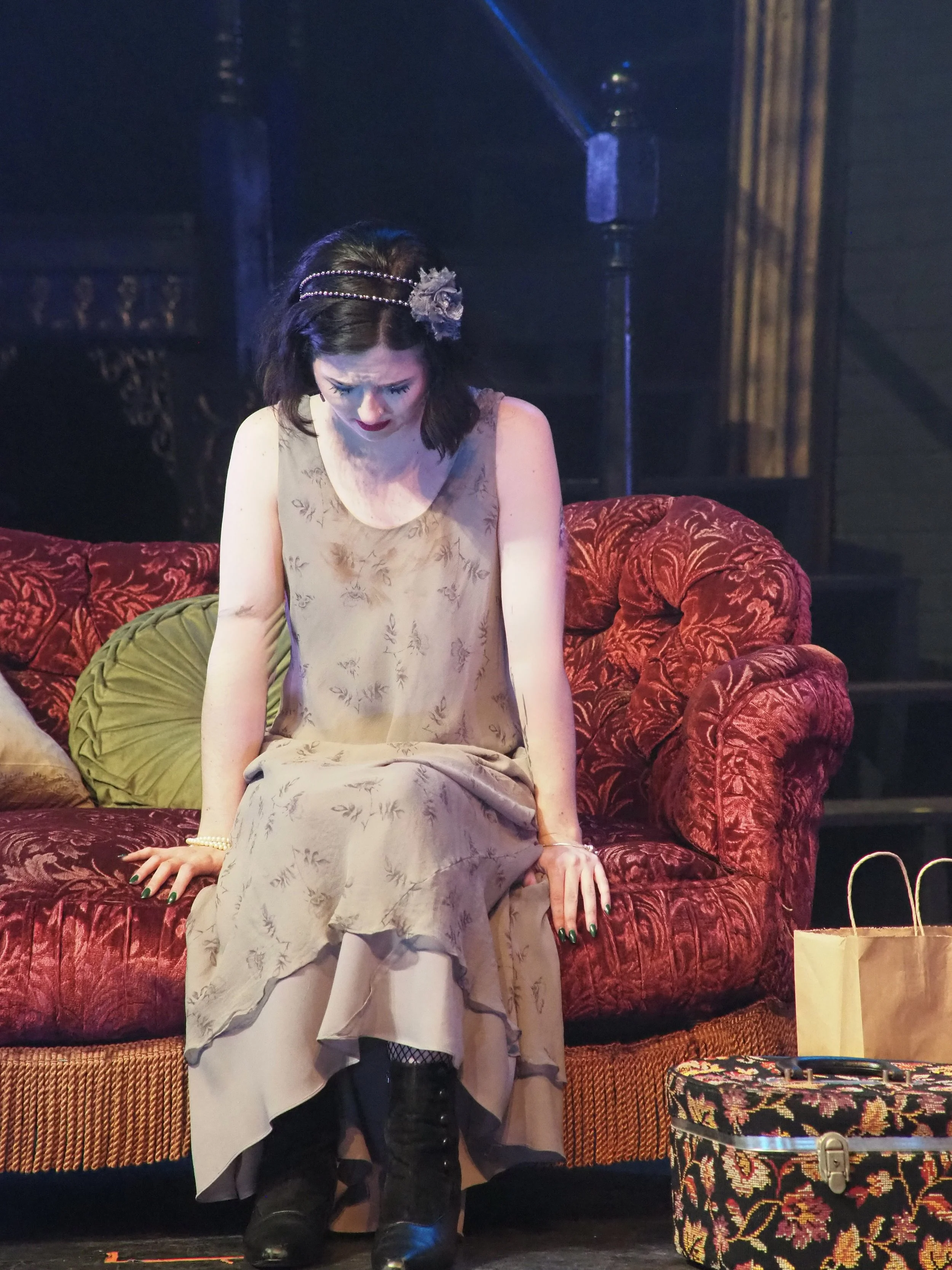 A woman sitting on a red vintage sofa, looking down with a somber expression, wearing a beige dress, black boots, and a floral headpiece, with shopping bags and a decorative box at her feet.