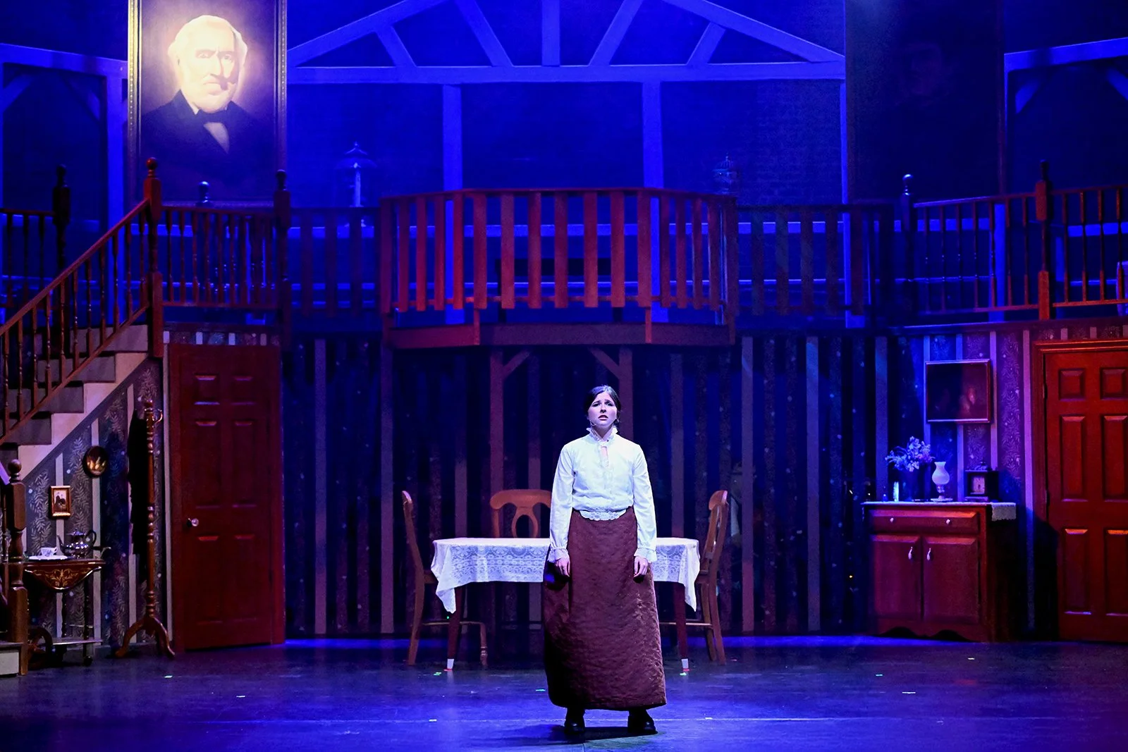 A woman standing alone on a theater stage with a set resembling a Victorian home interior, including a staircase, a table with a white tablecloth, and portraits on the wall, illuminated by blue lighting.