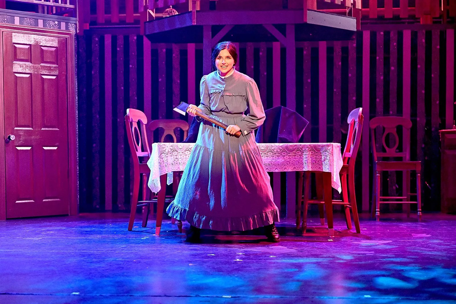 A woman in historical costume holding an axe on a stage with a wooden table, several chairs, and a staircase in the background, illuminated by colorful stage lighting.